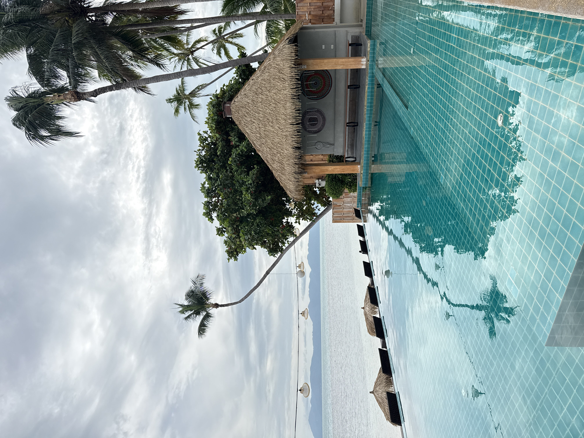 An infinity pool is in the foreground, its turquoise water reflecting the sky and palm trees. The pool is bordered by a tiled edge, and beyond it, several thatched umbrellas and lounge chairs are set up on a beach. In the background, a calm sea stretches to the horizon, meeting a mountainous coastline under a cloudy sky. To the right of the pool, a small, open-air structure with a thatched roof provides shade. It has two benches inside and is decorated with circular wall hangings. Palm trees and lush greenery surround the structure, and string lights hang across the scene, some illuminated. The location is likely a resort or beachfront property in the Ko Samui District of Thailand.