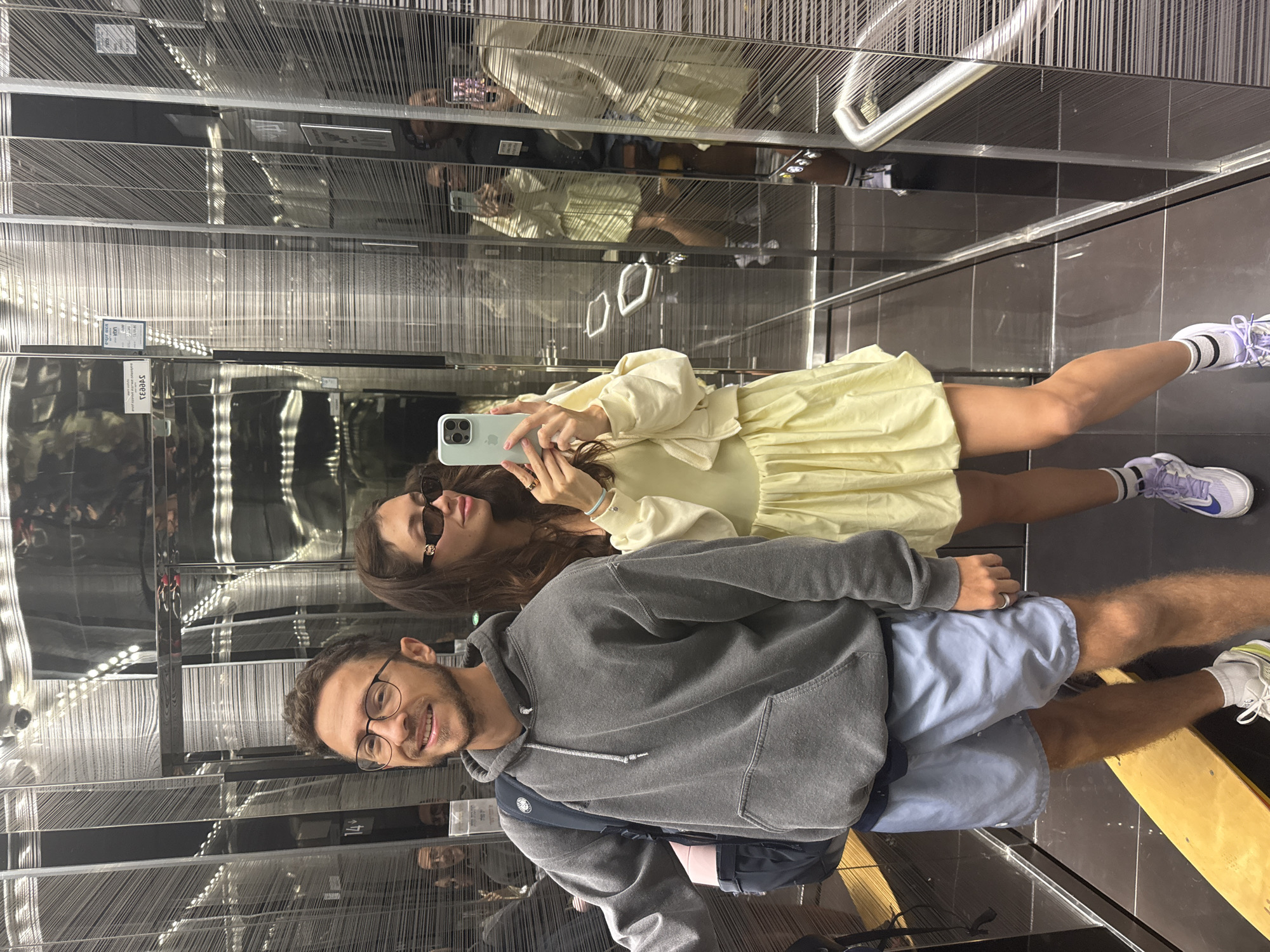 The photograph depicts a man and a woman in an elevator, both posing for a selfie. The man is wearing glasses, a gray hoodie, and blue shorts. The woman is wearing a yellow dress and sunglasses and is holding a light-colored smartphone. Reflections in the elevator's mirrored walls create multiple images of the individuals and the interior. The elevator's interior is decorated with a textured, striated pattern of thin, vertical lines. The floor of the elevator is dark-colored. A sign with the number "14" is visible on the elevator wall. The location is Tel-Aviv, Israel.