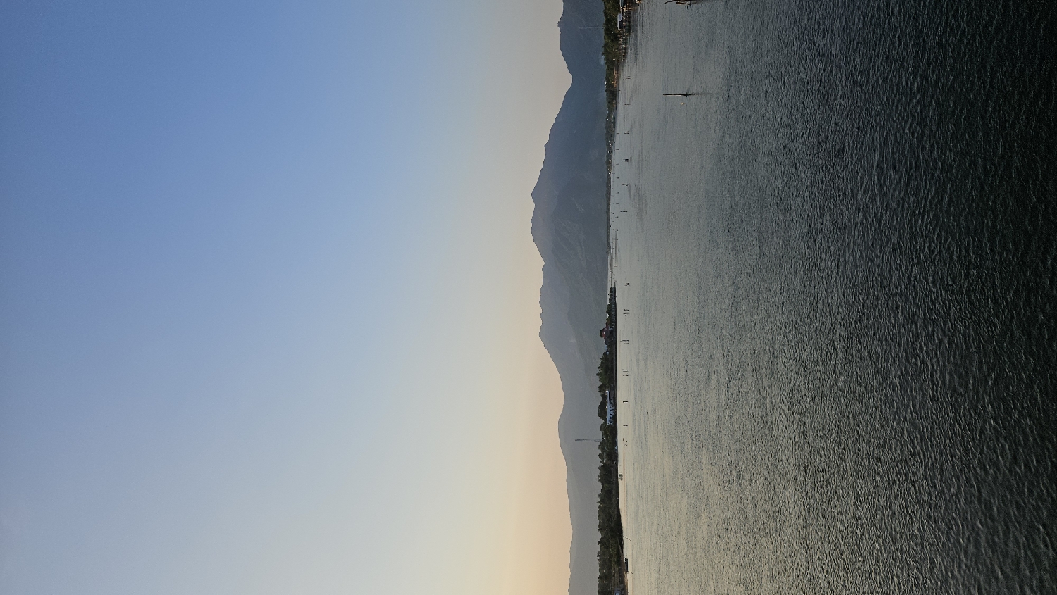 This tranquil landscape photograph, taken in Cam Đức, Vietnam, depicts a serene view of a wide body of water framed by distant mountains under a vast sky. The upper portion of the frame is dominated by a clear sky, transitioning from a medium blue at the top to a warm, pale yellow and orange near the horizon, characteristic of either dawn or dusk.
Below the sky, a long, hazy mountain range stretches horizontally across the middle distance, its undulating peaks appearing soft and indistinct due to atmospheric perspective. The lower half of the image features a calm expanse of water, gently rippling and reflecting the diffused light from the sky. Numerous slender, vertical poles, likely used for fishing or aquaculture, are sparsely distributed across the water's surface, particularly noticeable closer to the base of the mountains. Along the mountain's edge at the waterline, a few small, low-lying structures, possibly houses or fishing huts, are faintly visible.
No specific activity is explicitly depicted, lending a peaceful and calm mood to the scene. The weather appears clear with no visible clouds. No text is visible in the image.