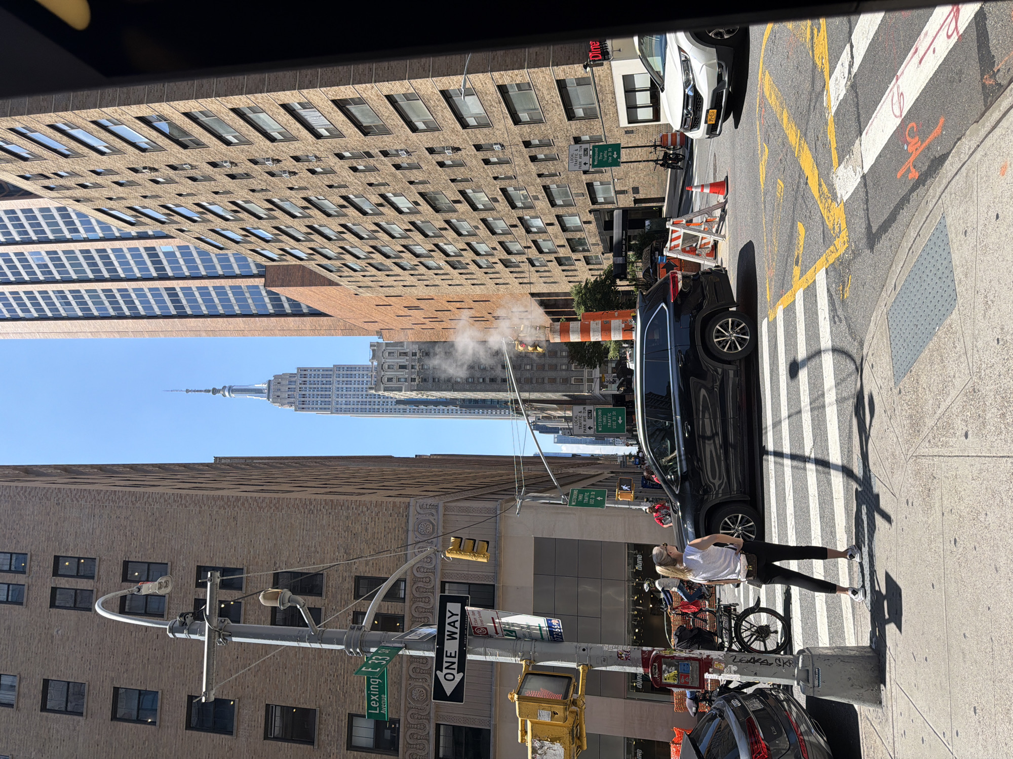 This image captures a street scene in New York City, likely during the daytime on a clear, sunny day. The perspective is from a vehicle, looking out at the urban environment.
The foreground features a pedestrian crossing with yellow and white painted lines. A black SUV is stopped at the crosswalk, partially obscuring a woman standing on the sidewalk. She is wearing a white t-shirt and black leggings, with blonde hair, and appears to be looking to the left. A bicycle is visible behind her. Further down the street, another car is parked.
The midground showcases a tall, brick building with many windows, characteristic of New York's architecture. Across the street, a steam plume rises from a street-level vent, suggesting an underground utility or building exhaust. Traffic cones and construction barriers indicate ongoing work. Several street signs are visible, including "ONE WAY," "Lexington Avenue," and directional signs for "WESTBOUND TRAFFIC USE 31 St."
In the background, the iconic Empire State Building stands tall against the blue sky, a clear indicator of the location. Other buildings of varying heights and architectural styles are also present, contributing to the dense urban landscape. The overall impression is a busy, typical day in Manhattan.