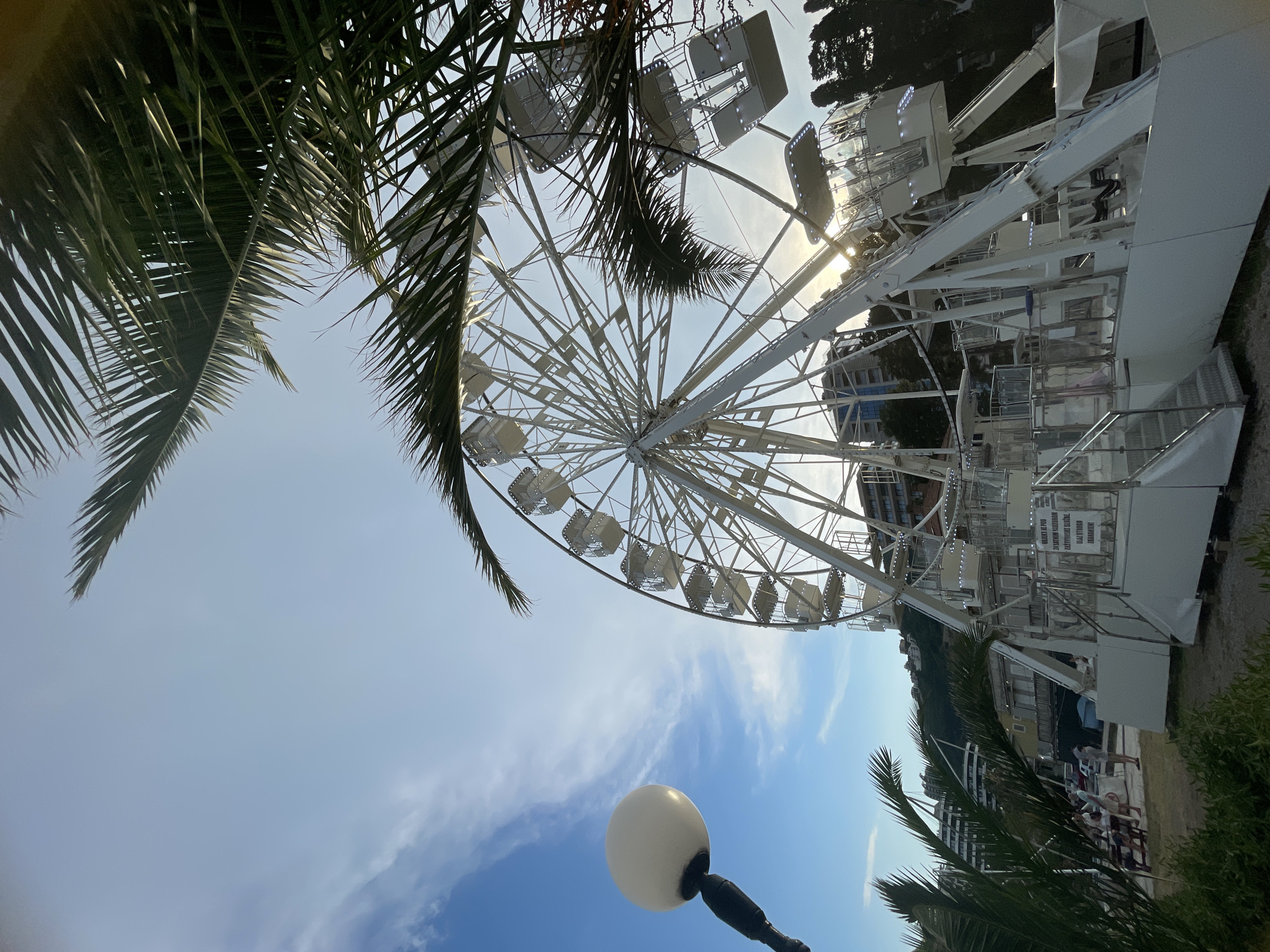 The image shows a large white Ferris wheel on a sunny day, possibly late afternoon due to the golden light. Palm fronds frame the top left and right of the image, suggesting a warm, tropical, or coastal location. The Ferris wheel has many enclosed cabins, and some are illuminated, indicating it may be open or about to open for operation. In the foreground, a white spherical lamp on a dark pole is visible. In the background, there are buildings and trees, with a hill in the distance. Some people can be seen near the base of the Ferris wheel, dressed casually, suggesting they are either visitors or staff. The sky is blue with some wispy white clouds. The overall atmosphere appears relaxed and leisurely, fitting for an amusement park or attraction. There is a sign on the side of the Ferris wheel structure with some text, but it is not clearly legible. The location is identified as Budva, Montenegro.