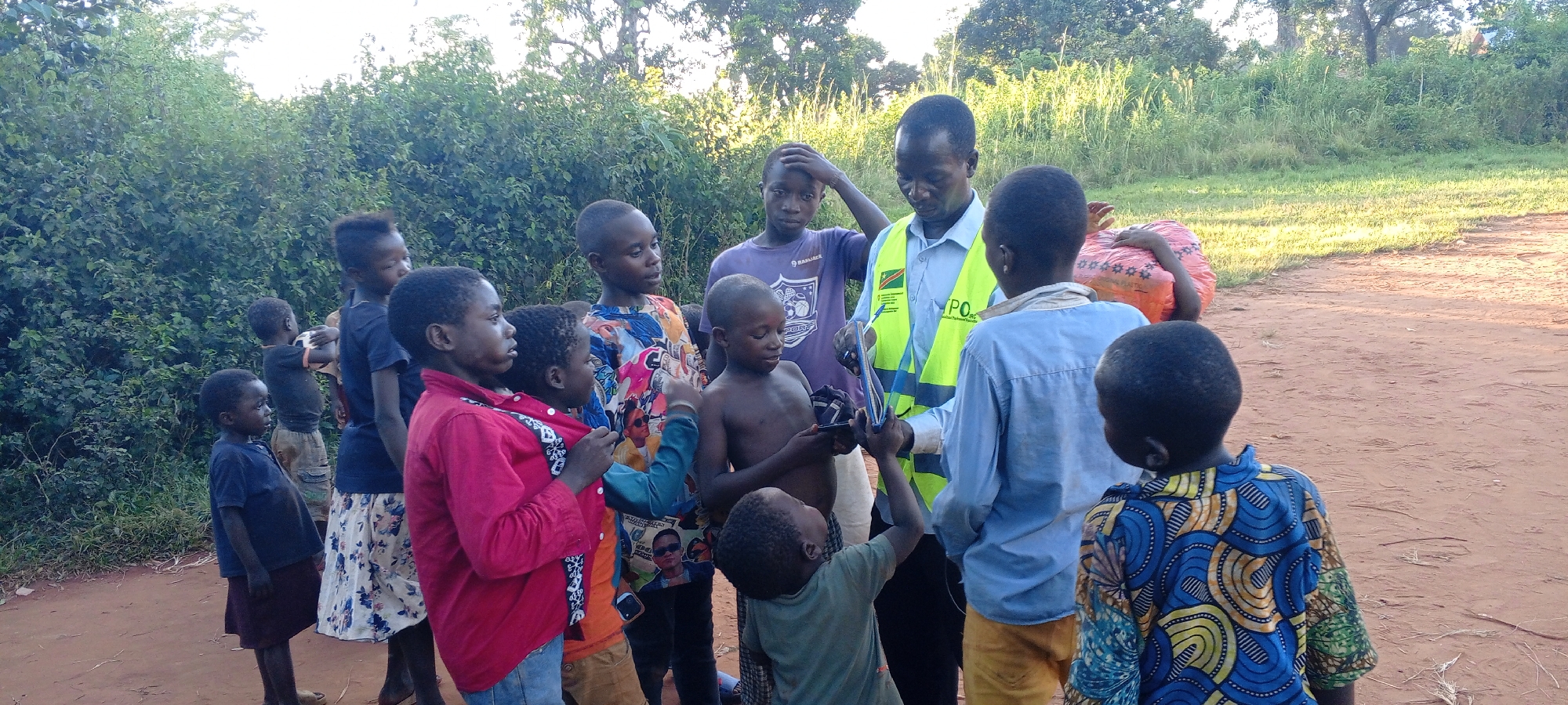 In Ngalula, Democratic Republic of the Congo, an adult male wearing a light blue shirt and a yellow safety vest is the central focus of a group of approximately thirteen children. The man appears to be showing something, possibly a phone or device, to the children. The children are of varying ages and are dressed in casual clothing. Some children are looking at the object in the man's hand, while others are looking at the man. The environment appears to be outdoors, with a dirt road or path visible underfoot. There is lush green foliage and trees in the background. A large red sack is on a child's back towards the background.