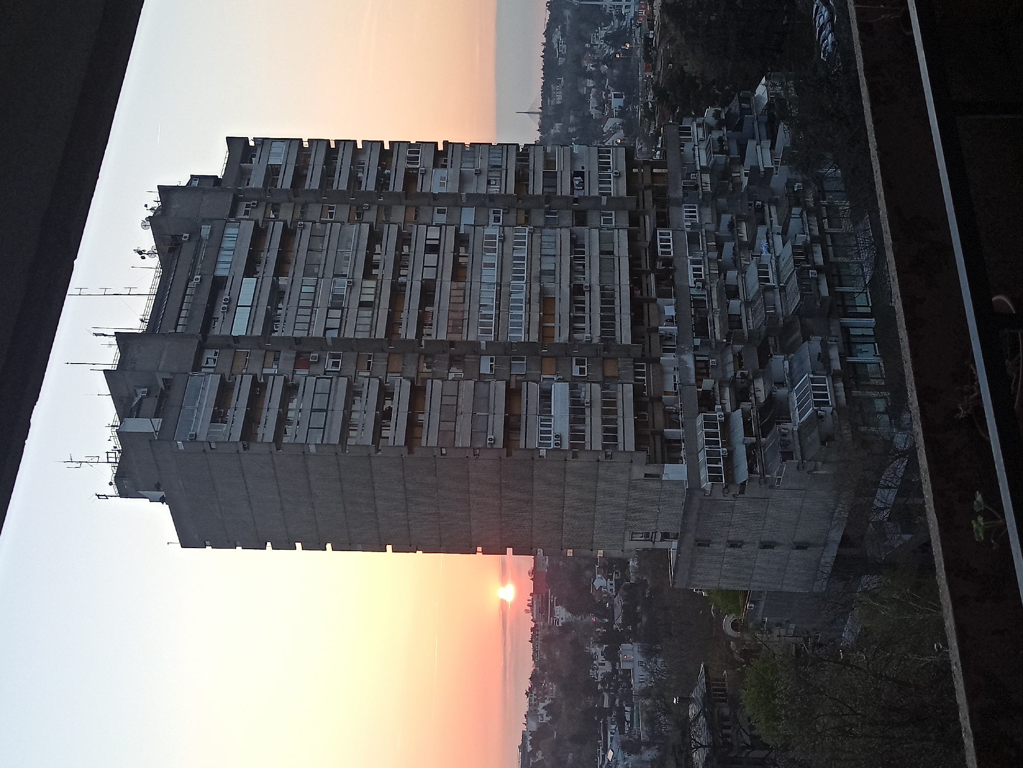 The image captures the Genex Tower, a notable Brutalist architecture landmark, in the Vozdovac Urban Municipality, Serbia. The building, a high-rise apartment block, is seen against the backdrop of a sunset. The tower's facade is marked by numerous windows and air conditioning units. The foreground is partially obscured by what appears to be part of a building or balcony from where the photo was taken. Below the tower, the image captures elements of the urban environment, including other buildings and vegetation, bathed in the light of the setting sun.