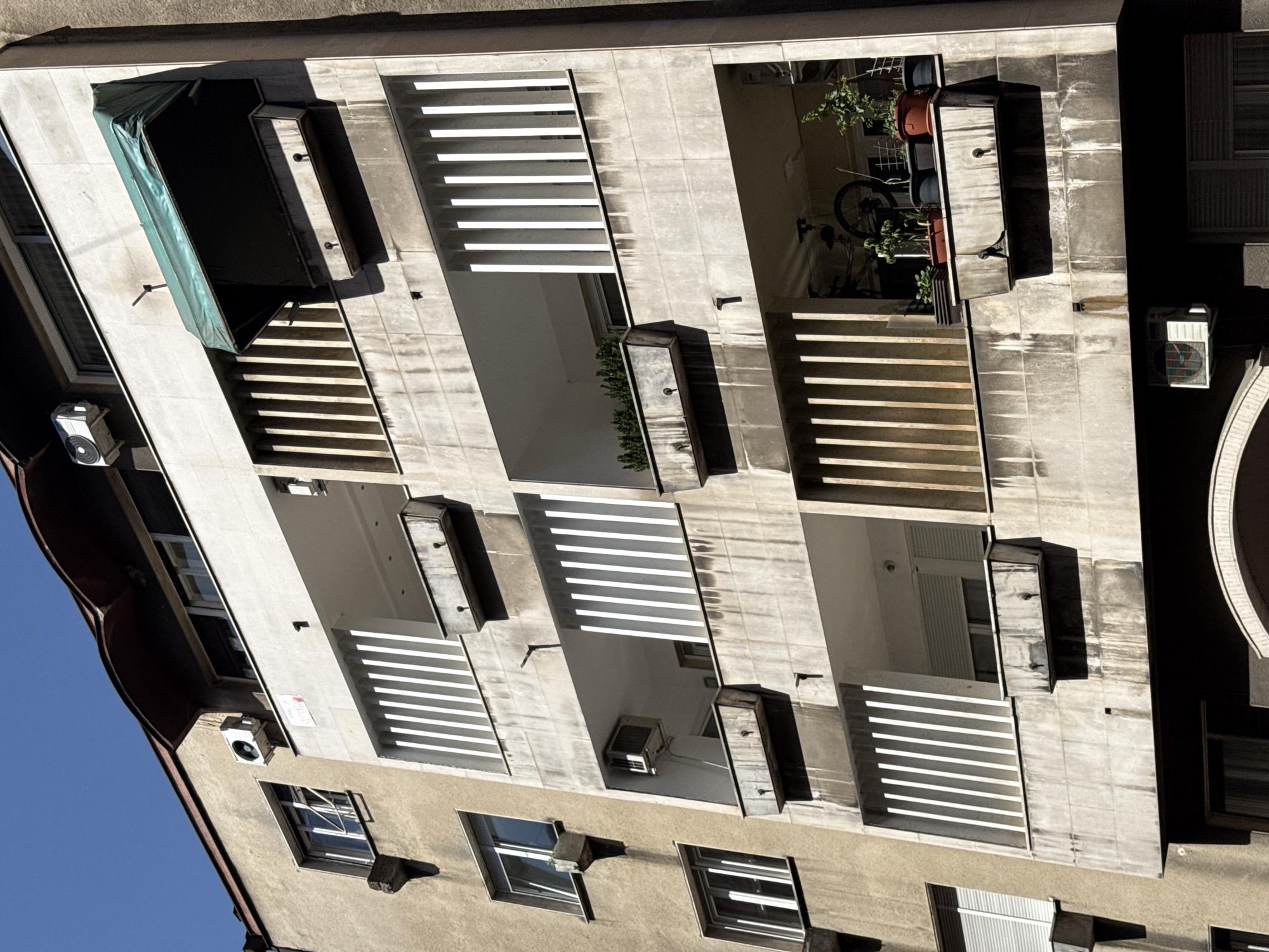 The image shows a multi-story building in Belgrade, Serbia. The architectural style features a concrete facade with balconies and horizontal slat sunshades. Some balconies contain plants and an air conditioning unit. The sky is visible in the background. Windows are visible on the lower floors. An awning is present on one balcony.