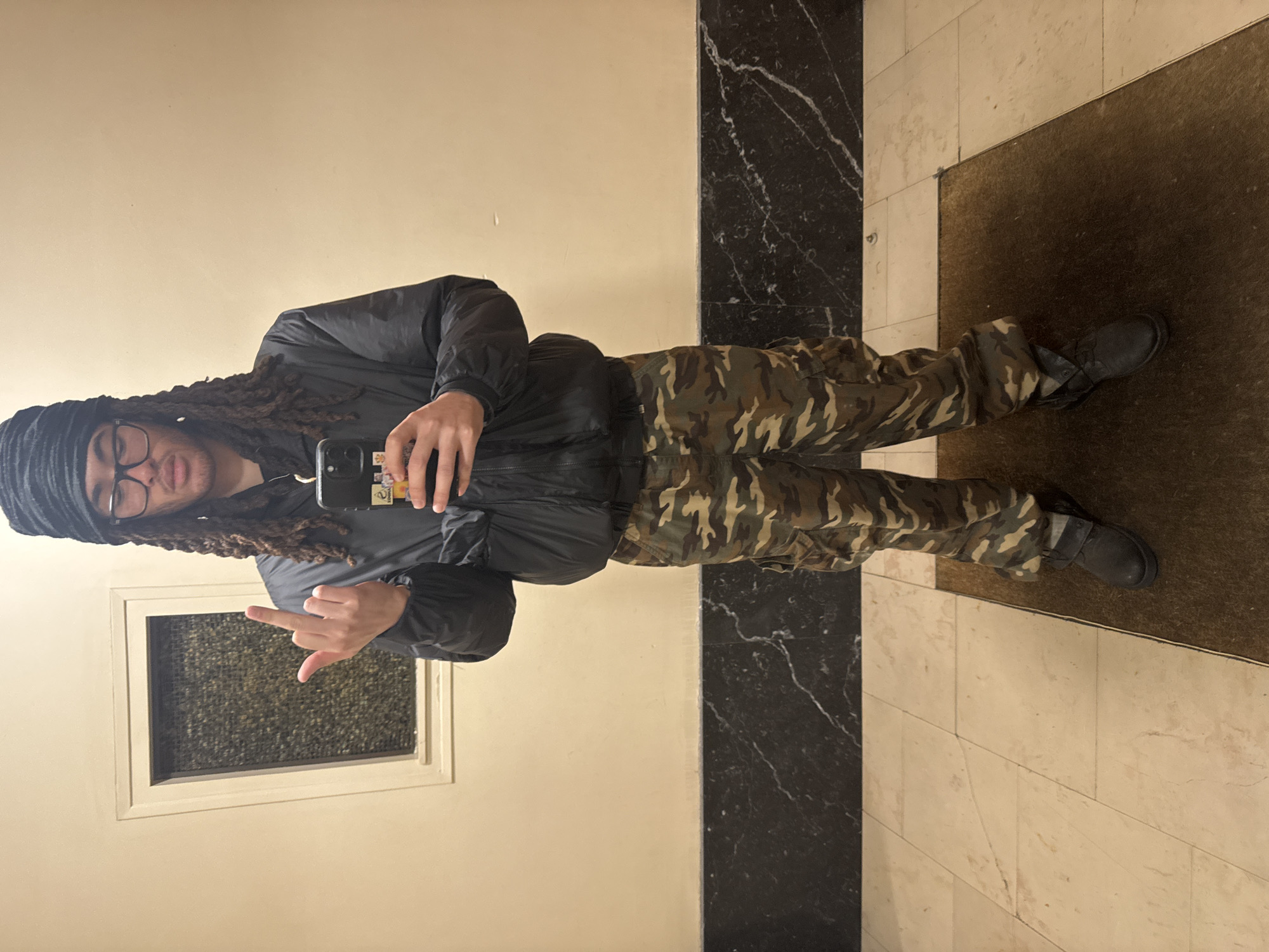 A person is standing indoors, taking a selfie in a hallway. They are wearing glasses, a beanie, a dark jacket, and camouflage pants. They are also wearing dark boots. The person is holding a phone in their right hand and extending their left middle finger. The background includes a door with a window, beige walls, and a marble and beige tiled floor. There is also a brown mat on the floor. The location is likely not in Paris, France, as there are no indicators of French architecture or design.