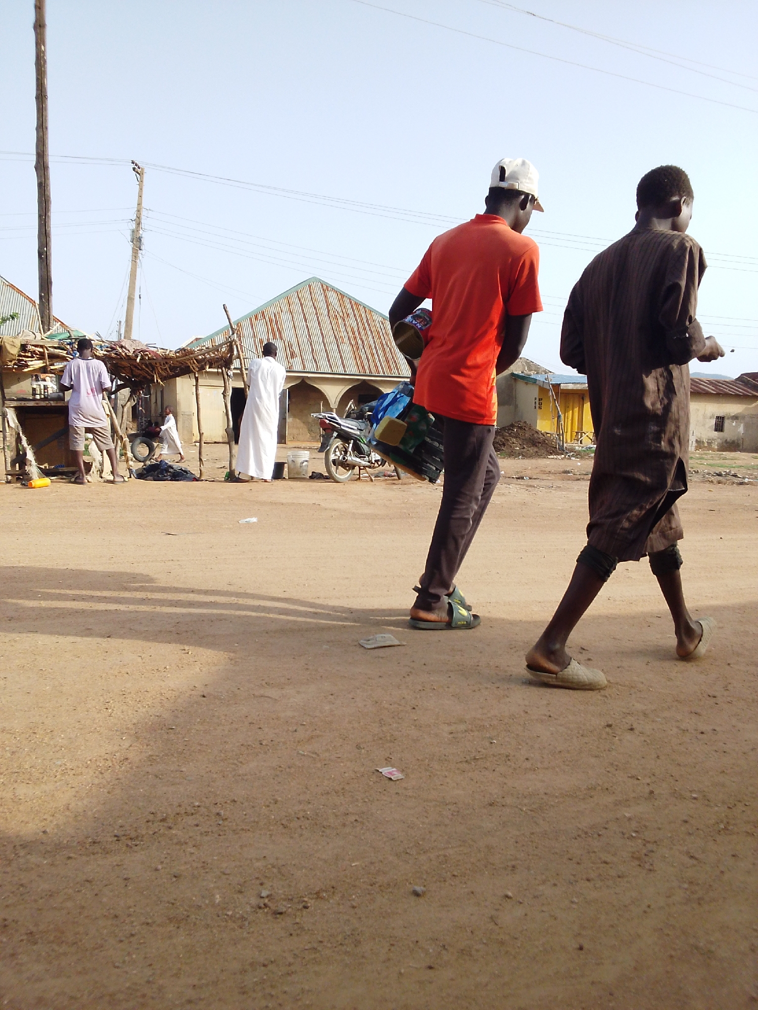 Two people walk right-to-left on a dirt road in what appears to be a residential neighborhood. The person on the left wears a bright orange shirt, dark pants, a white baseball cap, and sandals. He carries an unidentified item in his left hand. The person on the right wears a brown, long-sleeved garment over his legs, and light-colored sandals. In the background, there are buildings, including some with corrugated metal roofs, a motorcycle, and several people standing near a makeshift structure. Power lines and wooden poles are also visible. Based on the prompt, this is located in Bauchi, Nigeria.