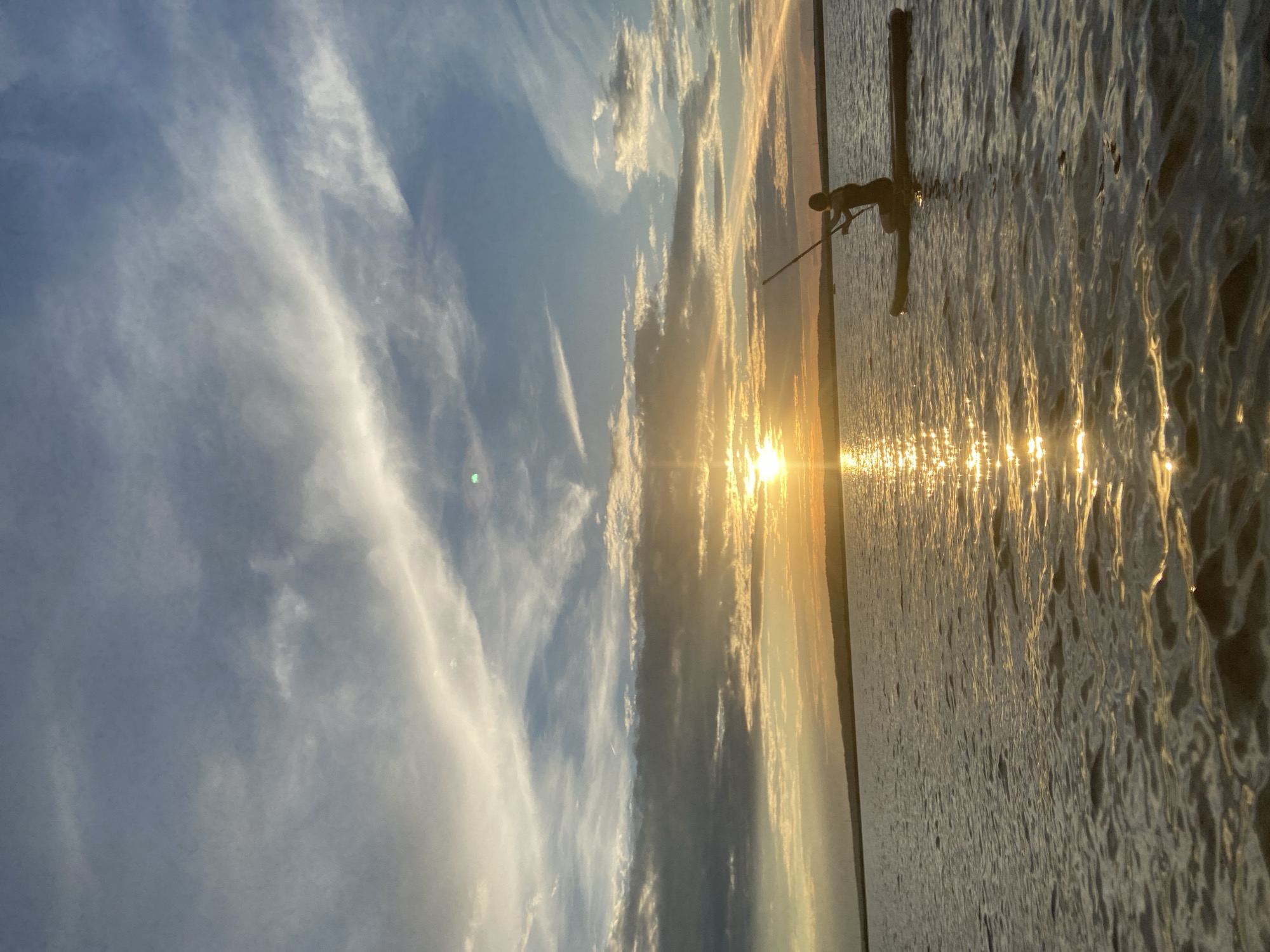 A person is paddleboarding on a body of water at sunset. The sun is low in the sky, casting a golden glow on the water and creating a reflection. The sky is filled with dramatic clouds, ranging from dark grays to bright whites. The person is silhouetted against the sunset, holding a paddle and appears to be in motion. The shoreline is visible in the distance as a dark, low line. The scene is serene and picturesque, capturing the beauty of a sunset over water. This scene is likely on the Kama River, as it is a prominent geographical feature in Chaikovsky, Russia.