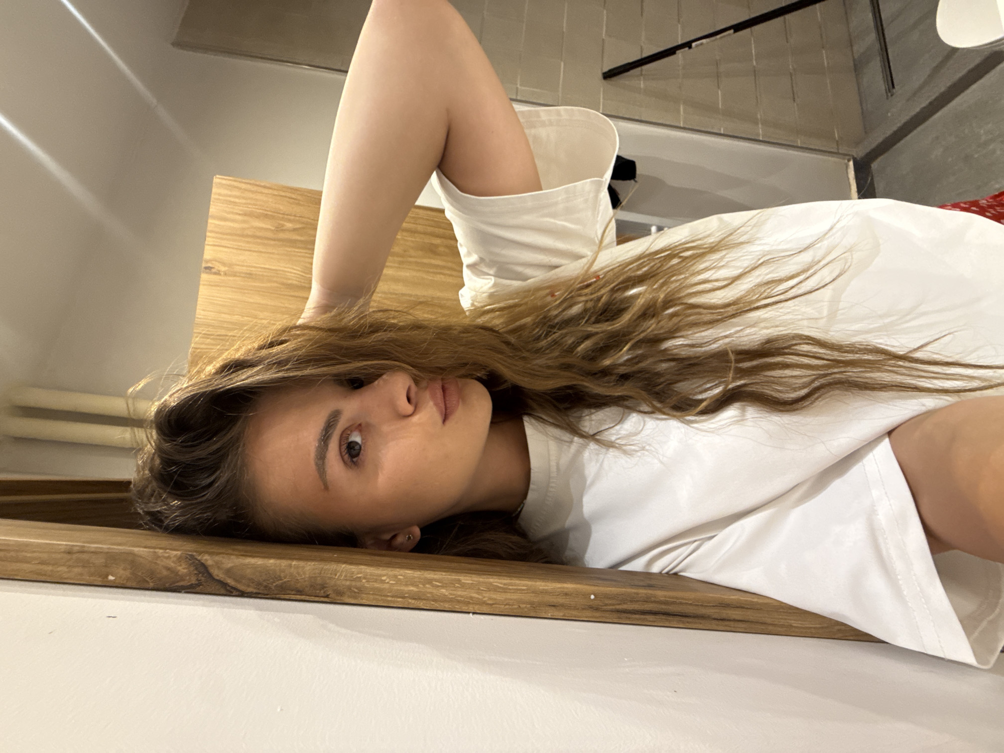 A woman is lying on a wooden surface, wearing a white T-shirt. She has long, wavy brown hair and is looking at the camera. The background is blurred, but there are hints of a room with white walls and a gray floor. The location is Unknown, Unknown.