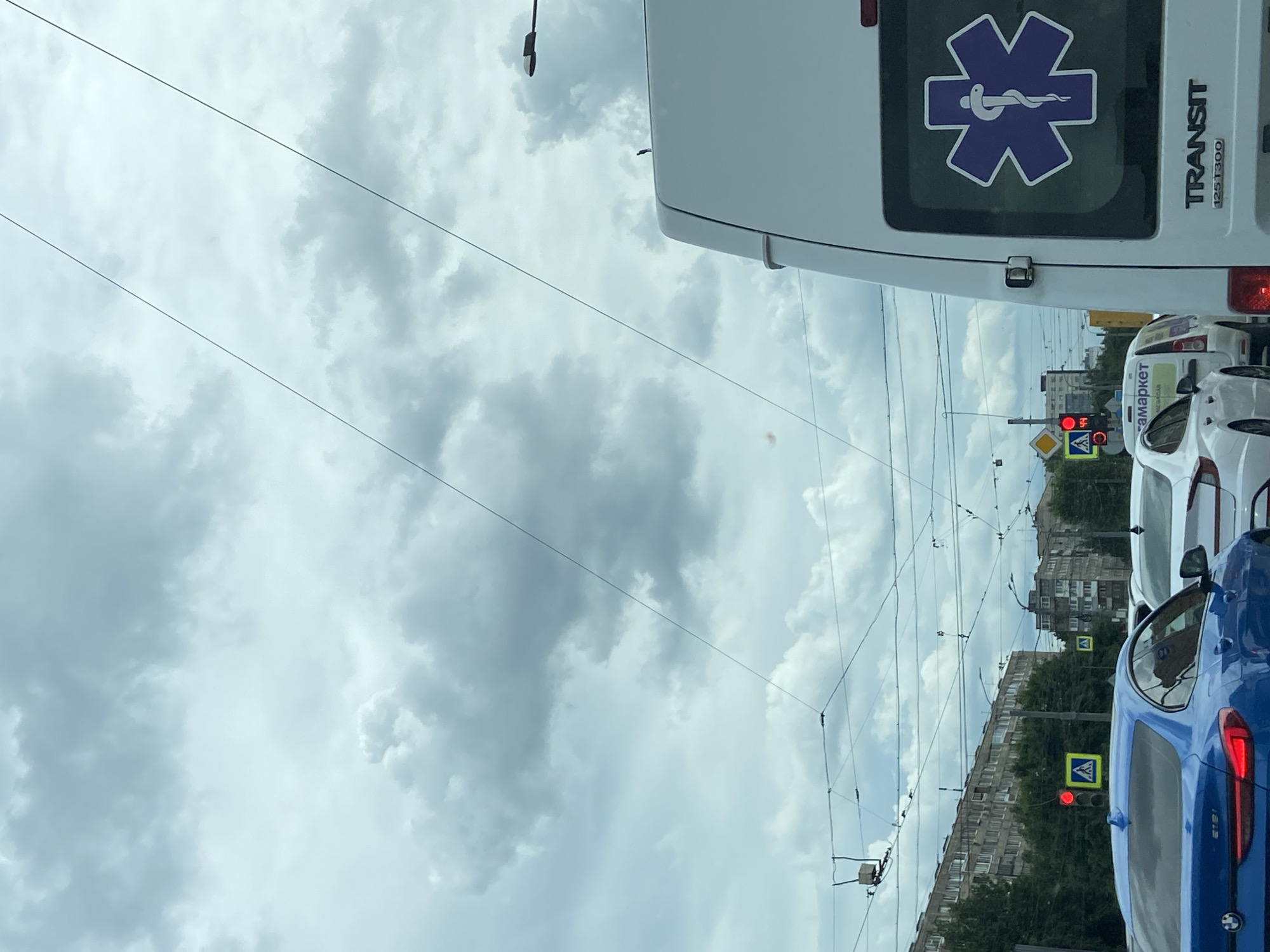 An overcast sky with heavy clouds looms over a busy city intersection. Traffic is backed up, with various vehicles visible, including a blue BMW in the foreground, a white sedan, and the rear of a white ambulance. The ambulance has a prominent purple Star of Life symbol on its rear window and the word "TRANSIT" printed below it. Traffic lights are visible, with one showing red. Buildings line the street in the background, appearing to be apartment blocks. Overhead, a complex network of wires crisscrosses the sky, likely for public transportation like trams. The scene suggests a typical urban environment experiencing traffic congestion.