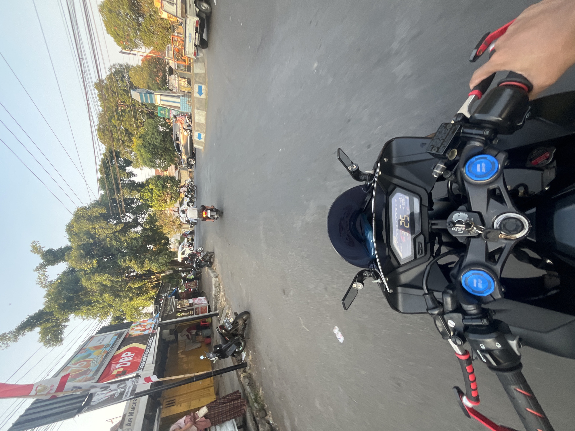 The image captures a first-person perspective of someone riding a motorcycle through a street in Pecangaan, Indonesia. The street is paved and appears to be a moderately busy thoroughfare. On the left side of the street, there are several storefronts, including one with a prominent red sign that says "DRP". A woman in traditional attire is visible near one of the shops. Several motorcycles and a few cars are present on the road, indicating ongoing traffic. In the distance, a roundabout is visible with a blue and white monument at its center, marked with blue arrows indicating traffic flow. Trees line the sides of the street, and power lines are strung overhead. The sky is clear and a light blue, suggesting it is daytime, likely late afternoon due to the soft lighting. The motorcycle's dashboard displays a speed of 25.