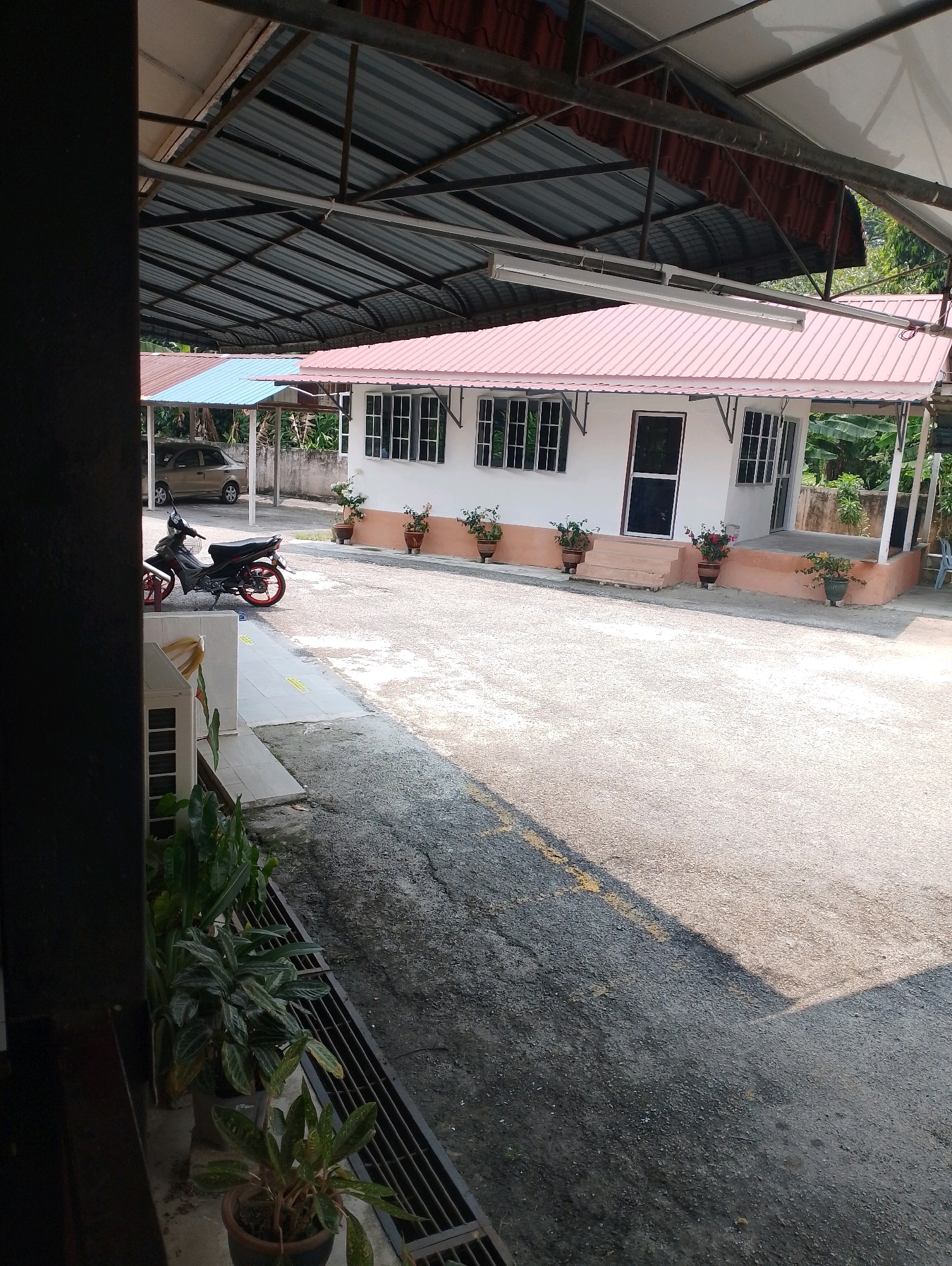 The image captures an outdoor scene in Pulau Tiga, Malaysia, focusing on a small building with a pink-tiled roof and a white facade. Several windows with white frames are visible on the building. In front of the building, there are potted plants placed along a coral-colored base. A concrete pathway leads up to a small set of steps and a door. To the left of the building, a motorcycle is parked on an asphalt surface. Beyond the building, there is a car parked under a separate canopy with a blue roof. The foreground features a dark, vertical structure, possibly a pillar or wall, and a row of potted plants. The ground is a mix of asphalt and gravel, with yellow markings visible on the asphalt. Lush green vegetation can be seen in the background, suggesting a tropical environment. The sky is not directly visible, but the lighting indicates it is daytime.