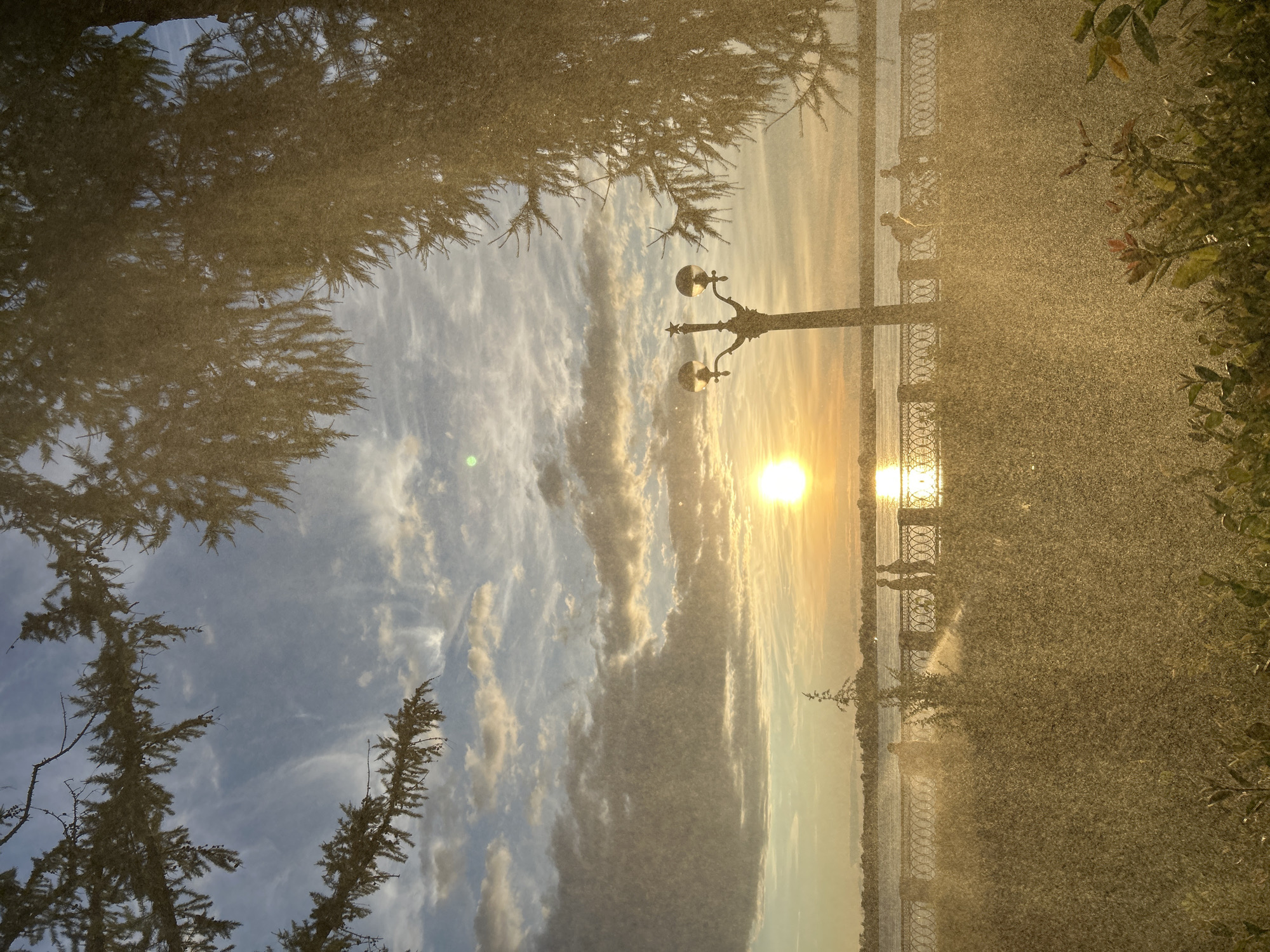 The image depicts a sunset scene at the Novokuybyshevsk embankment. The sun, a bright orb casting a golden reflection on the water, is partially obscured by clouds in the upper portion of the sky. The foreground is filled with a dense mist or spray, possibly from a sprinkler system, which creates a hazy, atmospheric effect. Silhouettes of people can be seen walking along the embankment, which is lined with an ornate metal railing. A lamppost with two globes stands near the railing. Evergreen branches hang into the frame from the top, adding a natural element to the urban landscape. The overall mood is serene and bathed in the warm light of the setting sun.