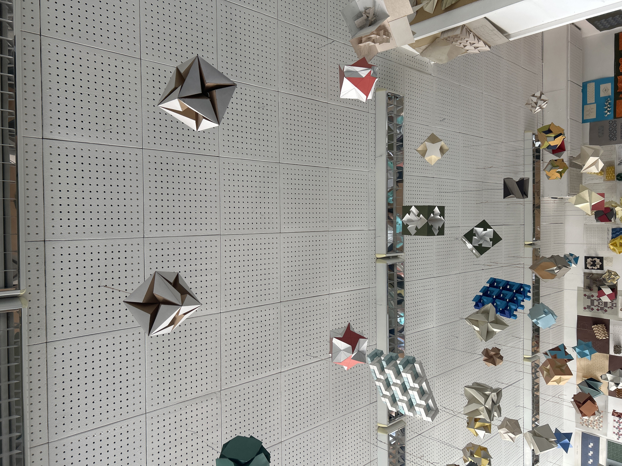The media file captures an indoor scene showcasing an intricate art installation composed of numerous geometric, folded paper or cardstock shapes. These multi-faceted objects, resembling polyhedra or modular origami, are suspended at various heights throughout the space, creating a dynamic three-dimensional display. The colors of the shapes vary, including shades of silver, grey, white, red, blue, green, brown, and gold.
The primary background is a large wall covered in light grey or white square panels, each uniformly perforated with small, circular holes. This textured wall extends across a significant portion of the image. Several tall, slender mirrored columns or structural beams are integrated into the space, reflecting parts of the room and the suspended geometric forms, adding depth and visual complexity.
On the far right side of the image, additional flat artworks, design samples, or graphic displays are mounted on a wall, showcasing various patterns and possibly textile or paper designs.
The scene depicts a static exhibition or art display, likely in a gallery, museum, or an architectural design studio in Sofia, Bulgaria. The lighting appears even and artificial, typical of an indoor exhibition space. No people are visible, and there is no legible text on the hanging objects or the perforated panels. The overall impression is one of precise craftsmanship and mathematical elegance in design.