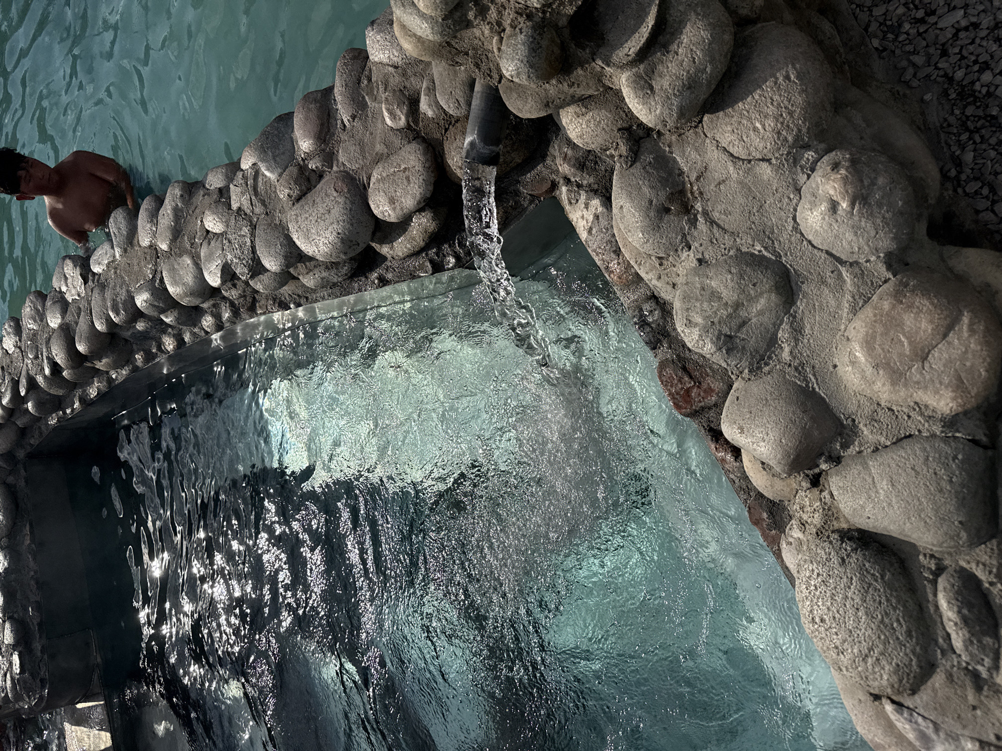 In Ak-Suu, Kyrgyzstan, an outdoor hot spring features clear, turquoise water with sunlight glinting off its surface. The pool's edge is lined with large, rounded stones, with a section of smooth, dark material visible on the left. A pipe extends from the rocky edge, discharging a steady stream of water into the pool, creating a cascade of bubbles and ripples. In the upper right portion of the image, a young person, shirtless, stands in the water, their body partially submerged. The background is out of focus, but appears to be an outdoor environment.
