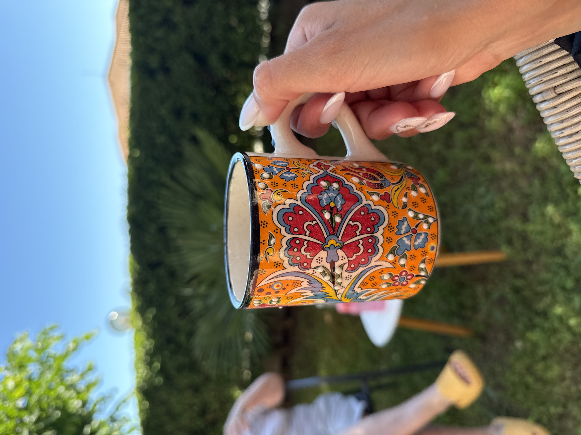 A close-up shot captures a hand holding an ornately decorated orange mug with floral patterns in red, blue, and yellow. The mug has a white handle and a black rim. The background is a soft-focus, sunny outdoor scene, featuring lush green foliage and a clear blue sky. To the left, a person's legs are visible, wearing yellow slides and sitting on a chair. In the mid-ground, a small white table with wooden legs is present. The scene appears to be set in the city of Kosharitsa, Bulgaria, given the architecture visible in the blurred background which suggests a Mediterranean climate. The overall impression is one of relaxed outdoor leisure.