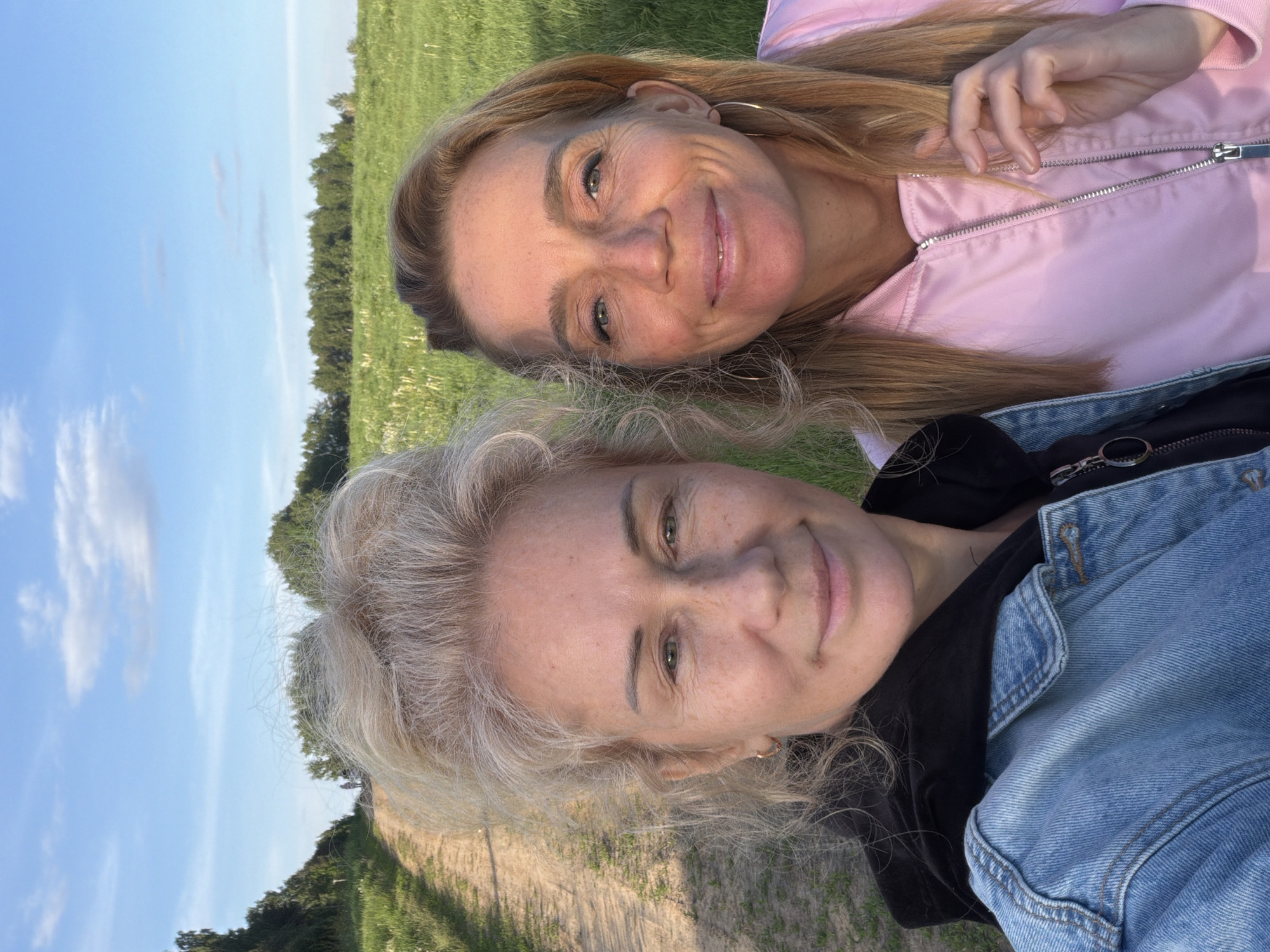 Two women are captured in a close-up, rotated outdoor portrait, likely a selfie, with their heads close together. The image is rotated 90 degrees counter-clockwise, making the sky appear to the left and the ground to the right.
The woman in the upper portion of the frame has long, straight, warm-toned blonde hair and is wearing a light pink zippered jacket. She has a subtle smile and is looking towards the camera, with light-colored eyes. Her hand is visible near her chest, seemingly resting on her jacket. She wears large hoop earrings.
Below her, the second woman has shorter, curly, cool-toned blonde hair. She is wearing a black top layered under a denim jacket. She also smiles faintly and looks directly at the camera with light-colored eyes.
The setting appears to be a rural or natural area, possibly in Норд Снт, Russia. In the background, there is a vibrant green field, transitioning to a line of trees in the distance, all under a clear blue sky with a few wispy clouds. The lighting suggests late afternoon or early evening, with a soft golden glow on their faces and hair. Both women appear relaxed and happy, sharing a friendly moment in the open air. There is no visible text in the image.