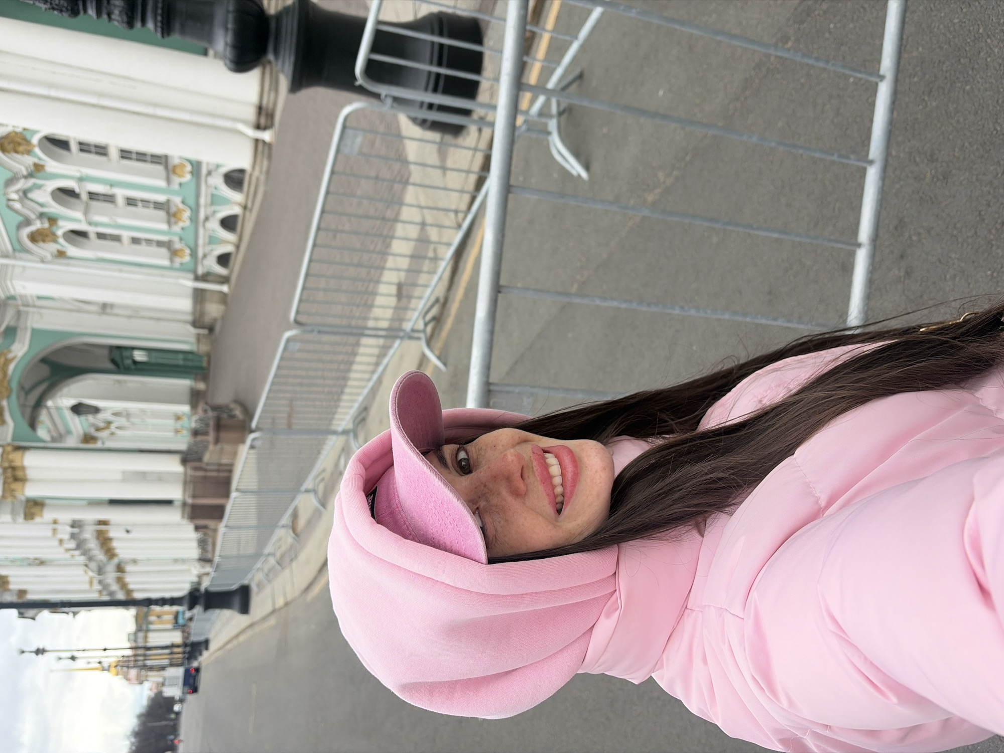 An outdoor portrait captures a young woman smiling brightly, looking slightly upwards and to her left. She has long, dark brown hair and prominent freckles on her cheeks and nose. She is wearing a light pink, puffy winter jacket and a matching pink hooded sweatshirt with the hood pulled up, partially covering a pink baseball cap. Her expression is joyful, with a wide smile revealing her teeth.
The background features a grand, ornate building characteristic of classical European architecture. Its walls are predominantly light cream or white, accented with intricate teal-green detailing and gold embellishments around arched windows and cornices. Several large windows are visible along the upper facade. In the midground, a series of light grey metal crowd barriers separate the woman from the building, suggesting a street or public area. A dark, possibly black, lamppost or pillar stands on the left. The scene suggests a city street or square. The sky appears overcast, indicating a daytime shot without direct sunlight. The woman's warm clothing suggests cool weather. No discernible text is clearly visible on any objects.
Based on the architectural style and context, the grand building is identifiable as part of the Hermitage Museum complex in Saint Petersburg, Russia.