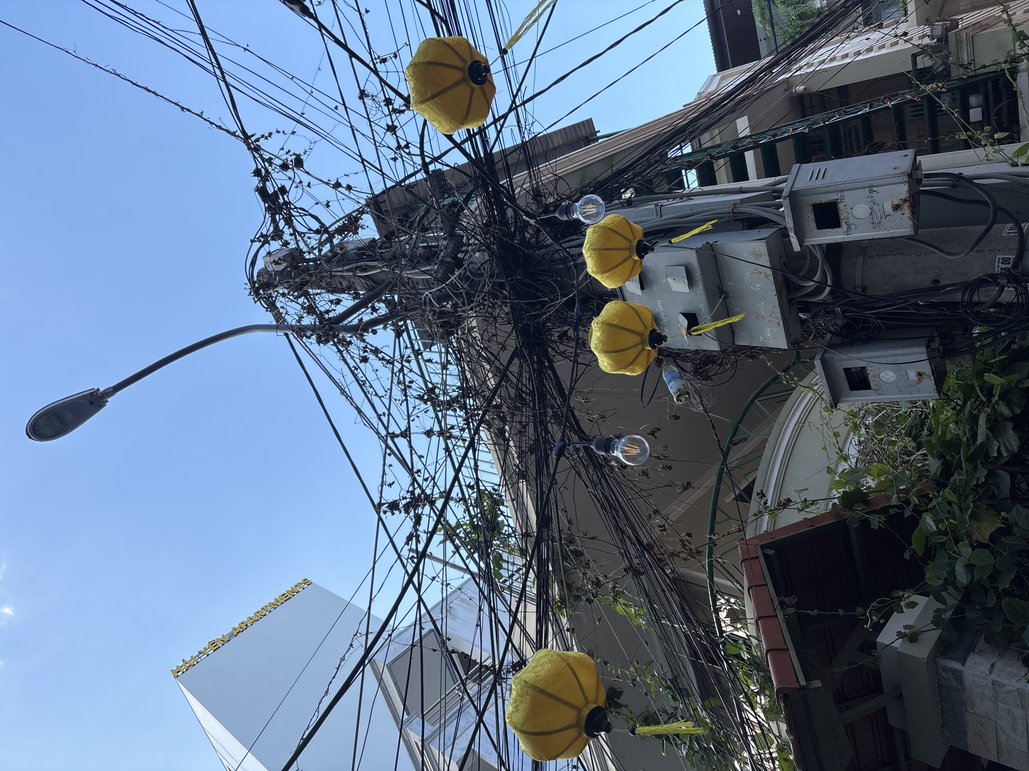 This media file captures a street scene in Nha Trang Ward, Vietnam, looking upwards towards a tangle of electrical wires against a clear blue sky. The wires are densely packed and appear disorganized, suggesting an urban environment with extensive utility infrastructure.
Several yellow, spherical lanterns are strung along the wires, adding a decorative element to the otherwise utilitarian scene. One of these lanterns is partially obscured by the wires. A bare incandescent light bulb is also visible, hanging from a wire.
In the background, buildings are partially visible. To the left, a modern, white building with golden text reading "HAVEN APARTMENTS" stands out. To the right, the facade of a building with traditional architectural elements, including a red-tiled roof and arched windows, can be seen. Overgrown green vines are creeping up the side of this building.
A street lamp arm extends into the frame from the left. The overall impression is of a bustling street with a mixture of modern and traditional architecture, heavily influenced by the overhead network of wires. The bright sky suggests it is daytime, likely with good weather. There are no people or animals visible in the frame. The scene conveys a sense of urban density and the visual complexity that can arise from extensive infrastructure.