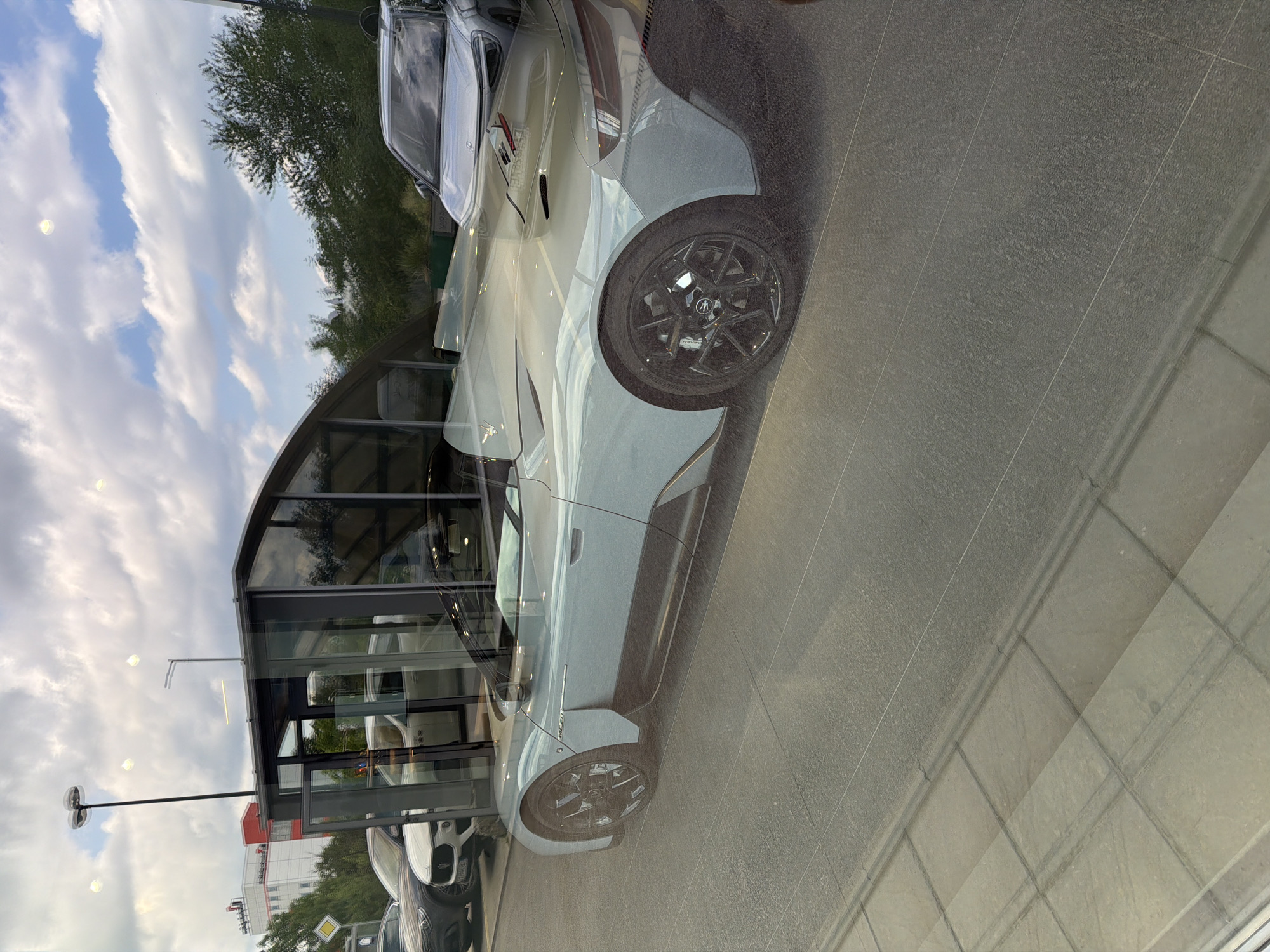 The image, originally rotated 90 degrees clockwise, captures a static scene through a large glass window of a car showroom, displaying vehicles while reflecting the outdoor environment.
The primary subject is a light-colored car, likely white or silver, visible in the central-lower part of the frame. It appears to be a Maserati, identified by the trident logo on its wheel's center cap and a faint logo on its side. Text "GT 2.0" is visible on the car's side panel, and the tire prominently displays "BRIDGESTONE POTENZA" along with "BRIDGESTONE" and a partial size, while the brake caliper clearly shows "BREMBO."
Dominating the scene are reflections on the glass window, which create a layered effect. These reflections include a partly cloudy blue sky, a dense mass of green trees, and various elements of an urban or suburban landscape. A tall street light with multiple lamps is reflected on the left side. Parts of other buildings are also reflected, including one with red accents. On the top right, a reflection of another vehicle, possibly a white car, is visible alongside a sign that faintly reads "TOYOTA," suggesting the presence of adjacent businesses or brands.
Inside the showroom, the floor is tiled in a light gray. Outside, the ground appears to be paved. The time of day is daytime, indicated by the visible sky and clouds. The scene conveys a clean, modern setting typical of a car dealership in Busmantsi, Bulgaria.