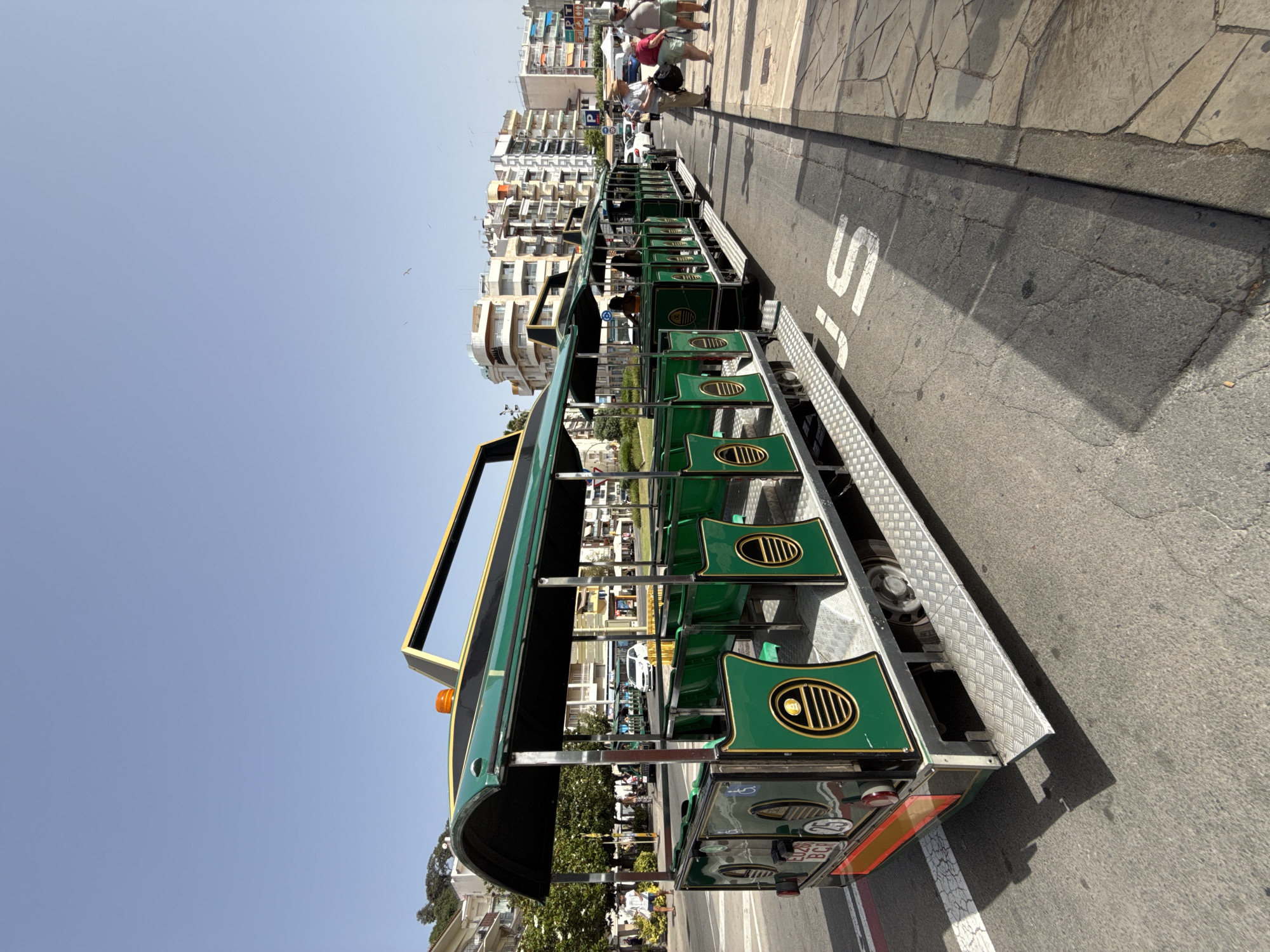 The image depicts a sunny day in Blanes, Spain, with a bright blue sky. A green tourist train, consisting of multiple open-air carriages with green seats and ornate gold detailing, is parked on an asphalt road. The train is positioned diagonally across the frame, with its front section visible on the left and its rear on the right. Several people are seen walking on a paved sidewalk beside the road, which is lined with apartment buildings and some trees. One building has a "P" parking sign. A white vehicle is visible in the background, behind the train. The road has white lines and a white "S" marking. A license plate on the back of the train reads "EL20 BCF". The overall scene suggests a typical tourist area, likely during daylight hours given the strong sunlight and shadows.