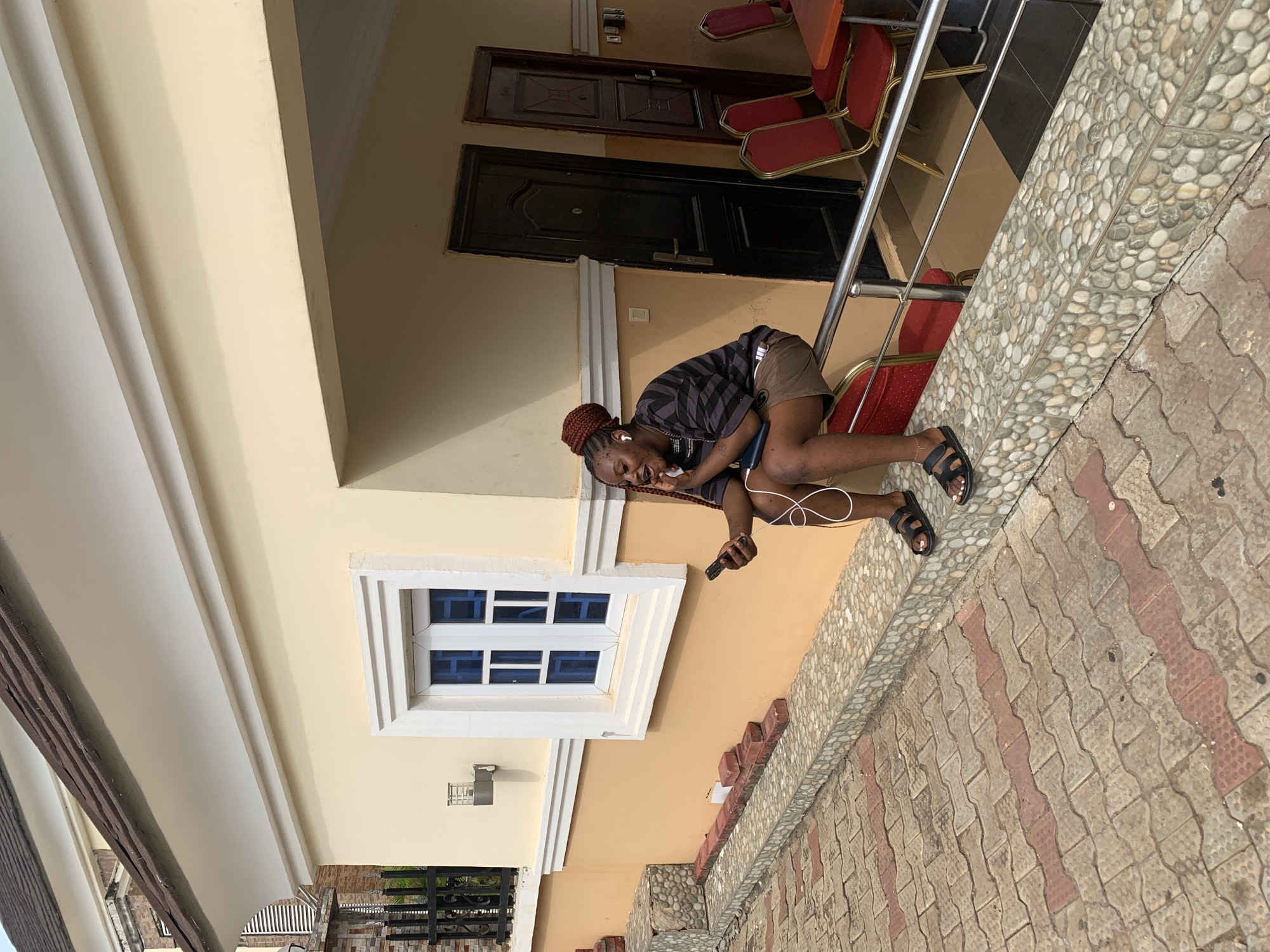 A young woman is squatting and smiling while using a mobile phone with headphones connected, outside a building. The building has a window with a white frame and blue glass, a metal balcony railing, and two dark-colored doors. The ground is paved with light grey and red-patterned bricks. The location could possibly be Lokoja, Nigeria, but without additional context, it's not possible to definitively confirm this.