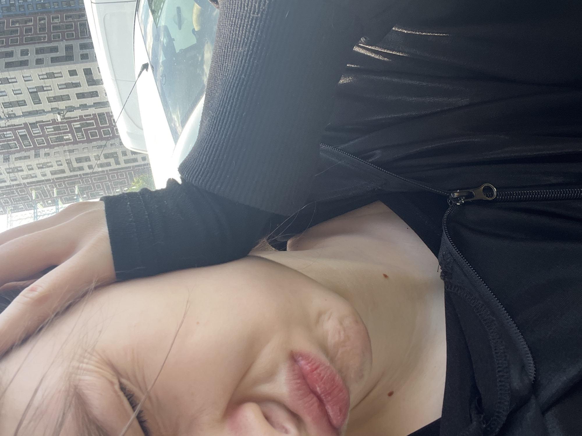 The image is oriented on its side, depicting a close-up selfie of a person, likely female, in what appears to be an urban setting, specified as Saint Petersburg, Russia. The person's face, including a partially visible eye, nose, and slightly parted, reddish-pink lips, occupies a significant portion of the foreground. Light brown or dark blonde hair strands fall across her face. Her skin tone is fair, and a few small moles are visible on her neck and upper chest area. One hand, wearing a dark long-sleeved garment, is resting near her head.
The person is dressed in a dark, likely black, zippered top or jacket. The sleeve and cuff area exhibit a ribbed texture, while the body of the garment appears to be a smoother fabric. A silver-toned zipper pull is clearly visible on the zipper, which runs along the side of her torso.
In the background, a white vehicle, possibly a car or van, is partially visible. Reflected in its windshield are inverted geometric patterns of numerous windows from adjacent urban buildings, indicating a dense city environment. The bright ambient light suggests it is daytime with clear weather conditions. The person's expression appears neutral or slightly contemplative. There is no visible text in the media file.