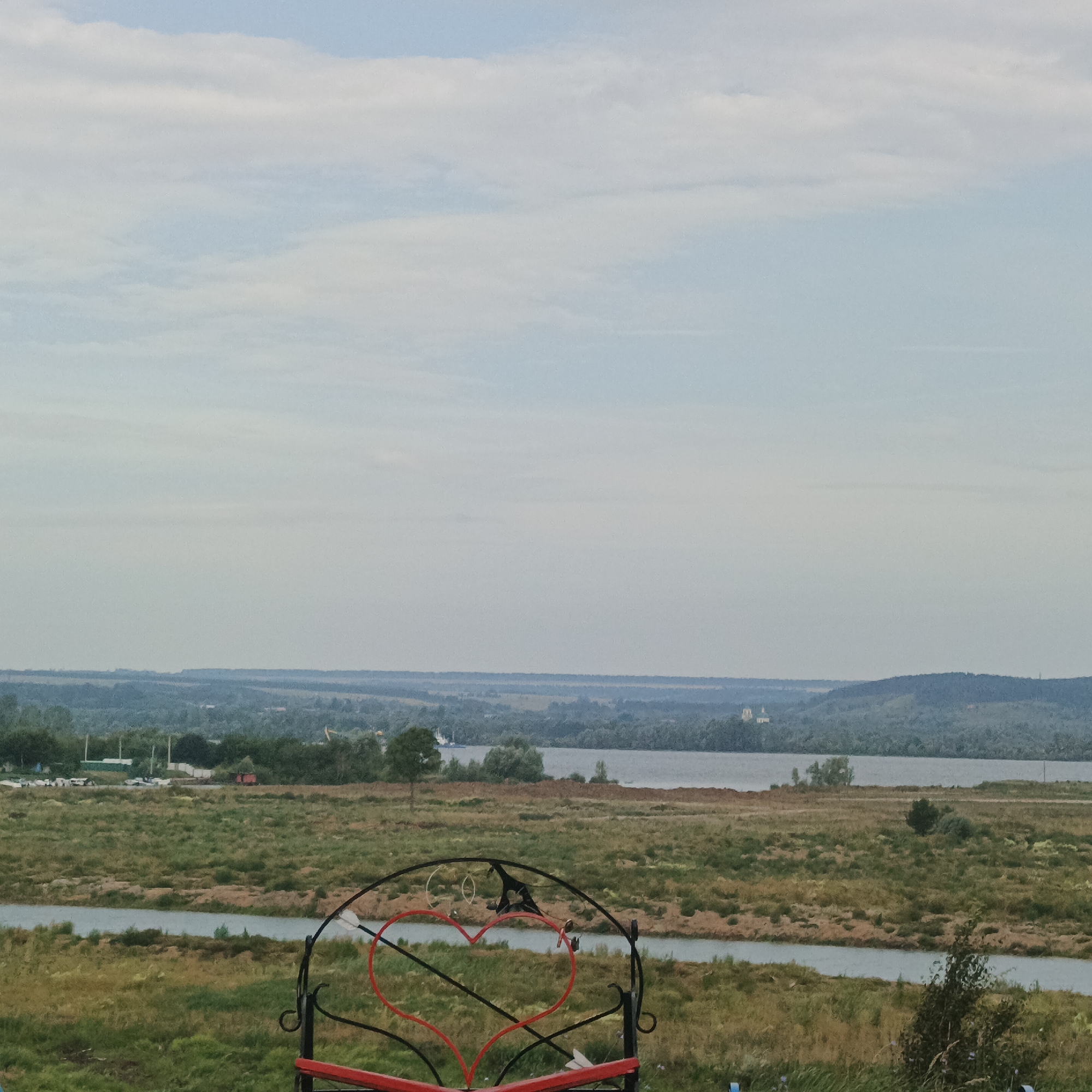 The image depicts a scenic overlook near Nikolo-Beryozovka, Russia. In the foreground, a decorative metal structure with red heart shapes is visible. Beyond this, a wide river flows through a landscape of grassy fields and scattered trees. In the distance, rolling hills covered in greenery recede into the horizon under a hazy, overcast sky. A few buildings and a church spire are faintly visible on the far bank of the river. The overall impression is one of a peaceful rural environment.