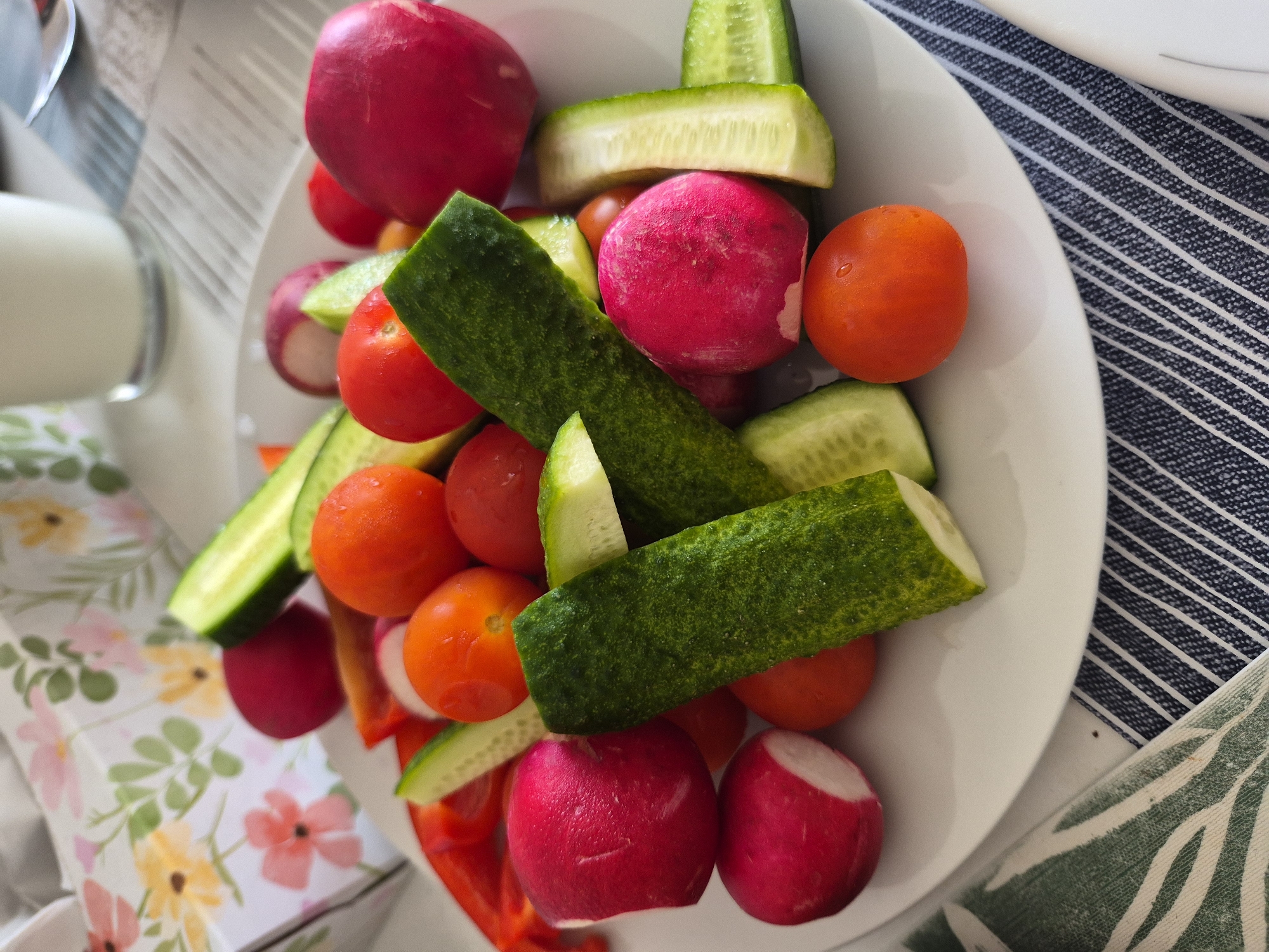 A white plate holds a variety of chopped fresh vegetables, including radishes, cherry tomatoes, and cucumber slices. The vegetables are arranged loosely on the plate, with some pieces overlapping. The plate is positioned on a table with a patterned placemat underneath. In the background, a glass of white liquid, possibly milk or yogurt, is visible, along with a floral-patterned box. The lighting suggests an indoor setting, likely a kitchen or dining area. No people or specific actions are depicted.