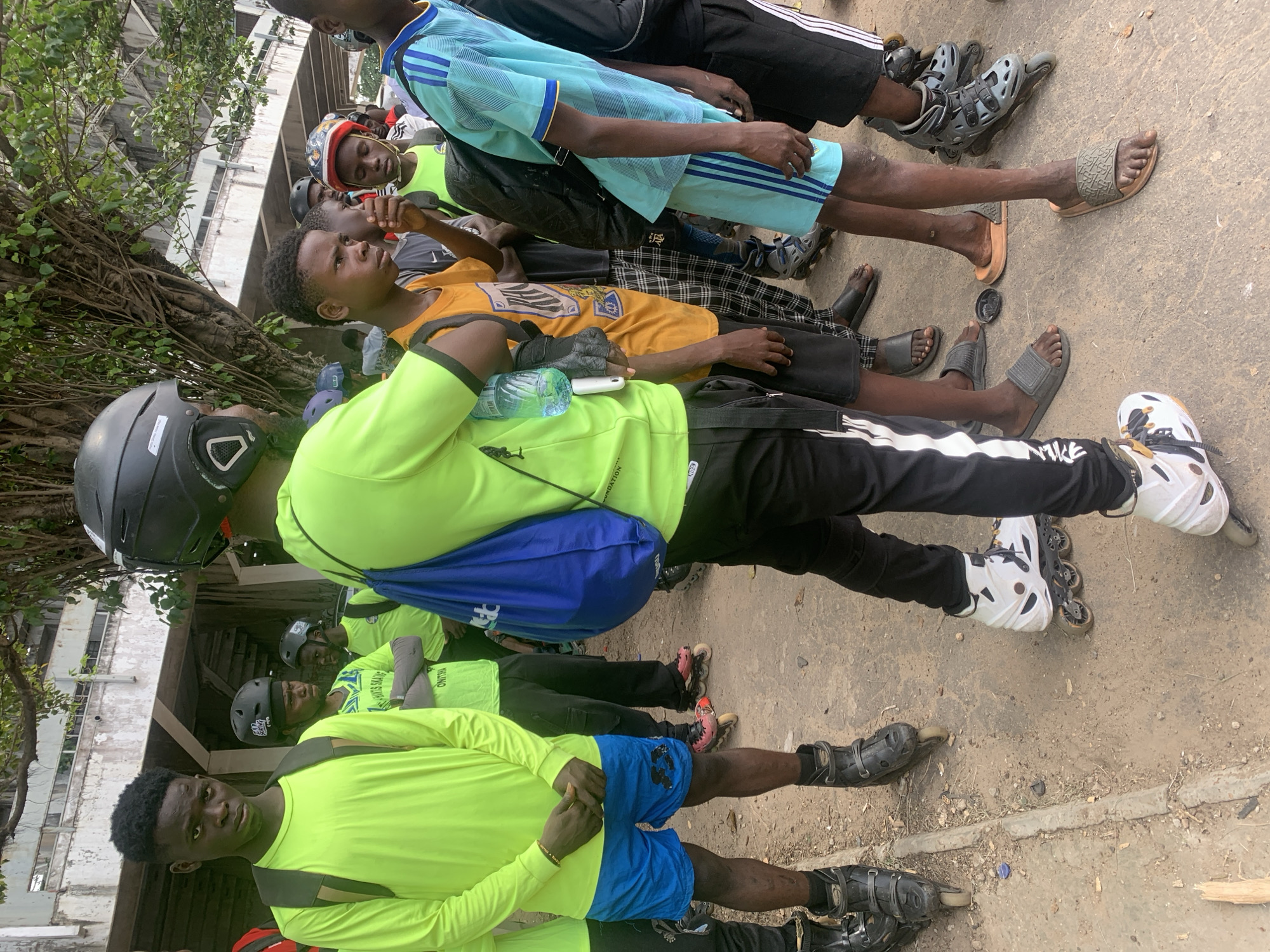 The image shows a group of young people, mostly boys, gathered outdoors on a dusty, paved surface. They are wearing casual clothing, including t-shirts, shorts, and pants, and many are wearing helmets, indicating they might be engaged in or about to engage in an activity like rollerblading or skateboarding. Some individuals are wearing rollerblades. The background features a large tree with dense foliage and a concrete structure that appears to be part of an overpass or building. The overall atmosphere seems informal, with the group appearing to be socializing or preparing for an activity. This scene is located in Alagbede, Nigeria.