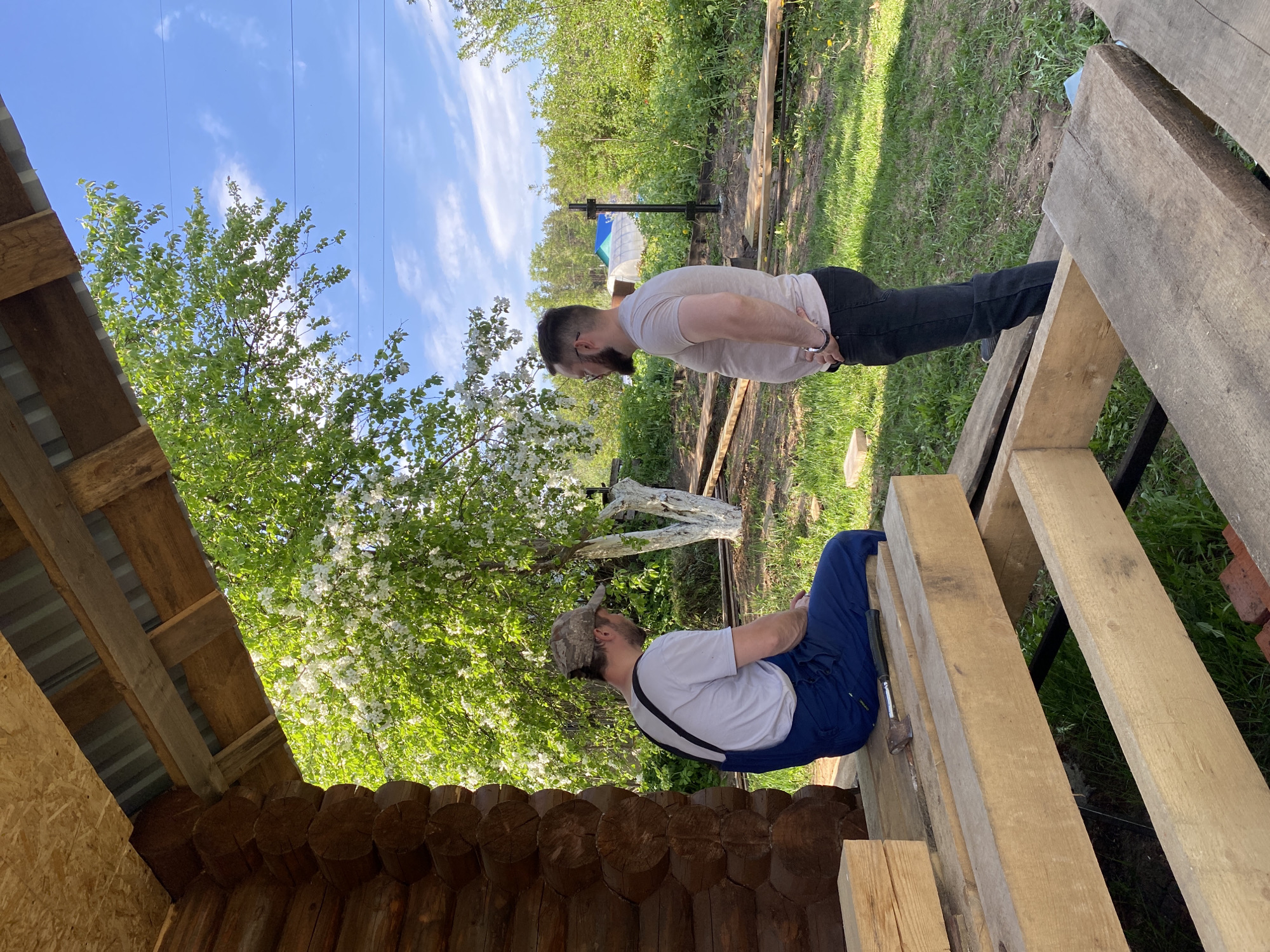 The image depicts an outdoor scene in Chaikovsky, Russia, featuring two men near a wooden structure under construction. One man is seated on wooden planks, wearing a camouflage cap, white shirt, and blue overalls with suspenders. An axe rests near him on the wood. The other man, standing and facing the seated man, is wearing a light-colored shirt and dark pants. In the background, there's grass, trees, a blue sky with scattered clouds, and a hint of a greenhouse. The wooden structure is constructed of logs and lumber with a corrugated metal roof. Overhead electrical wires are visible.