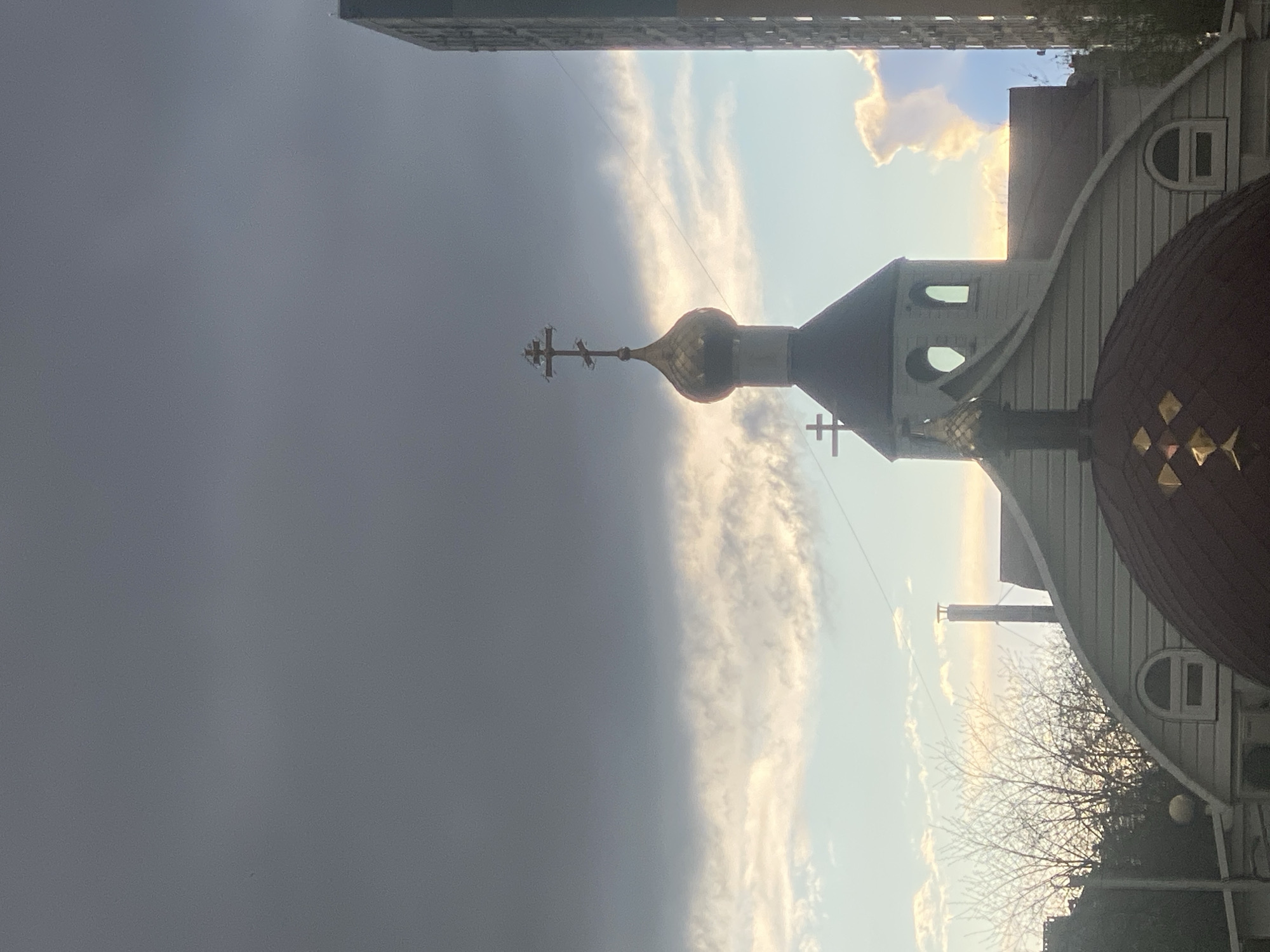 The image depicts an orthodox church in Unknown, Unknown, as viewed from a low angle. The church features a golden dome topped with a cross. The building facade has white siding and arched windows. The sky is divided between dark, stormy clouds and clear blue sky. A tall building is partially visible in the upper portion of the image. Bare tree branches are seen near the base of the church structure.