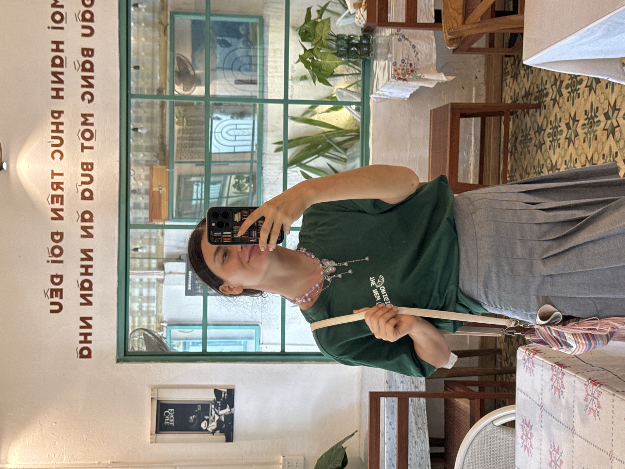 A young woman with dark hair pulled back stands in what appears to be a cafe, taking a selfie in a mirror. She is wearing a dark green t-shirt and a grey pleated skirt. A light-colored crossbody bag with colorful tassels hangs from her right shoulder. She is also wearing a delicate necklace with a butterfly pendant. In the background, there is a wall with Vietnamese writing, a large window with green trim, and several tables and chairs, suggesting a cafe environment. The writing on the wall, which is reflected in the mirror, is: "Đời là những trang đường. Dừng lại để ngắm, để yêu, để hy vọng, để cống hiến." This translates to: "Life is a journey of pages. Stop to admire, to love, to hope, to dedicate." The overall setting is brightly lit, with plants visible through the window and decorative elements on the tables. The presence of Vietnamese writing strongly suggests the location is within Vietnam, and more specifically, potentially in Phu Thuy Ward, given the context provided.