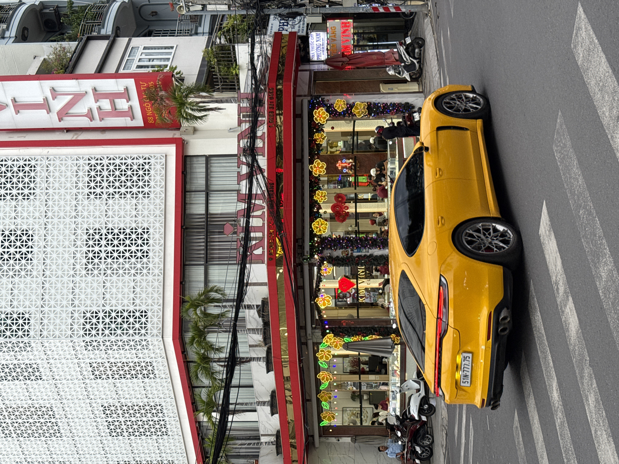 A yellow Porsche Panamera is stopped at a pedestrian crossing on a city street in Nha Trang, Vietnam. The car is facing away from the viewer, its rear visible. The license plate reads "51N-777.75". In the background, a building with a distinctive white lattice facade and a red awning is visible. The awning is decorated with yellow floral lights and garland. A sign above the awning reads "KIM VINH". Further up the building, a large vertical sign displays the letters "VINH" in red. Several palm trees are planted in pots along the second-floor balcony. Motorcycles are parked on the sidewalk to the left of the building. People are visible inside the store, appearing to be customers and staff. The street is paved with asphalt, and the pedestrian crossing is marked with white stripes. The sky is overcast, suggesting a cloudy day. The overall scene depicts a commercial street in Nha Trang.