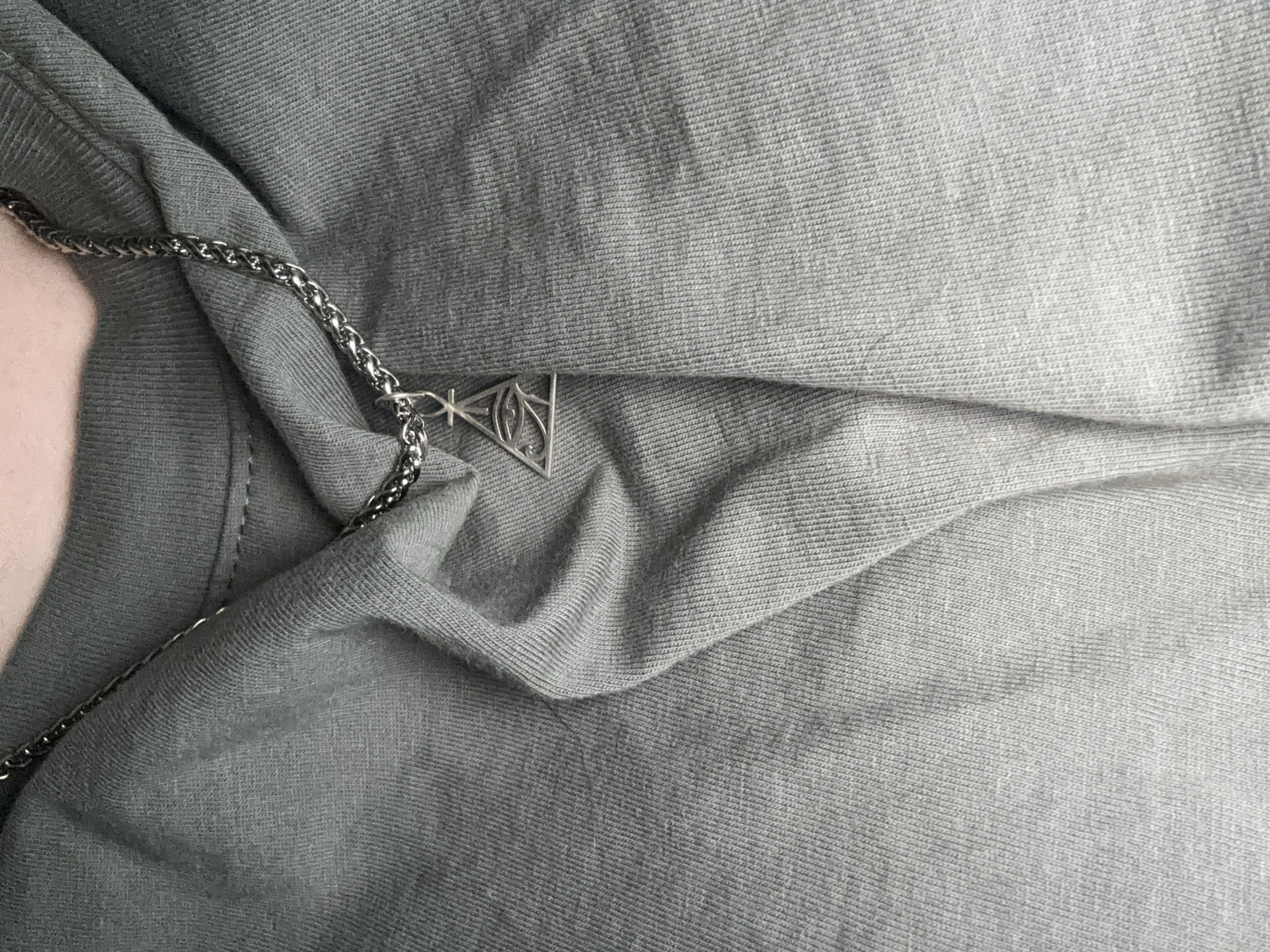 The image shows a close-up view of a person's neck and chest wearing a grey t-shirt. A silver chain necklace with a triangular pendant featuring an eye is visible. The lighting is soft, and the background is out of focus, highlighting the necklace and the texture of the t-shirt. The image does not contain any elements that would indicate a specific location in Saint Petersburg, Russia.