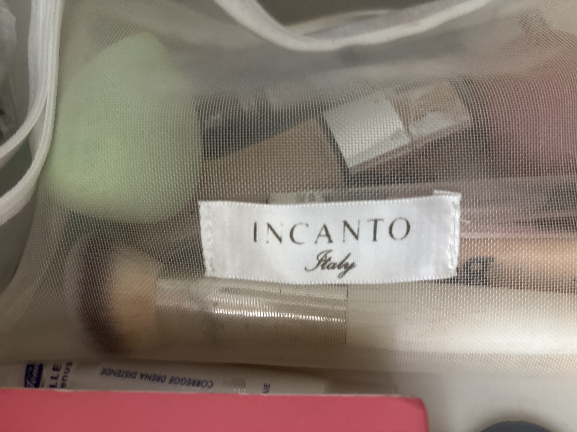 The image shows a close-up, eye-level view of a makeup bag's contents, seen through a translucent mesh material. The bag contains various makeup items, including what appear to be foundation bottles, makeup sponges, and possibly brushes. A white tag is visible with the text "INCANTO Italy" printed in black. To the left of the tag, another white label with blue text reads "LLe enus" and "CORREGGE DRENA DISTENDE". The overall environment appears to be indoors, with soft lighting. There are no people or specific identifiable landmarks visible in the image, nor any indication of the city of Ldzaa, Abkhazia. The focus is entirely on the contents of the makeup bag.