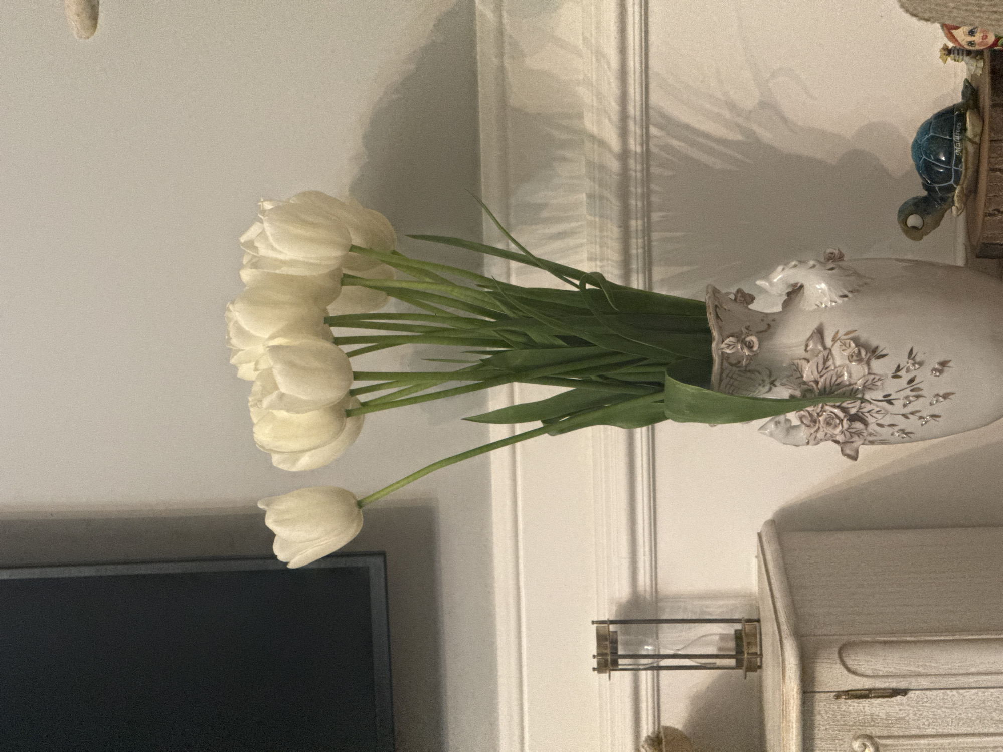 A vase of white tulips sits on a white cabinet in a room with white walls. A television is visible in the lower left of the frame. The vase is decorated with raised floral designs. A small decorative turtle figure is visible on the cabinet, next to the vase. The setting is indoors, but the image does not contain information to ascertain that the location is in Dzoraghbyur, Armenia. No people are visible in the image.
