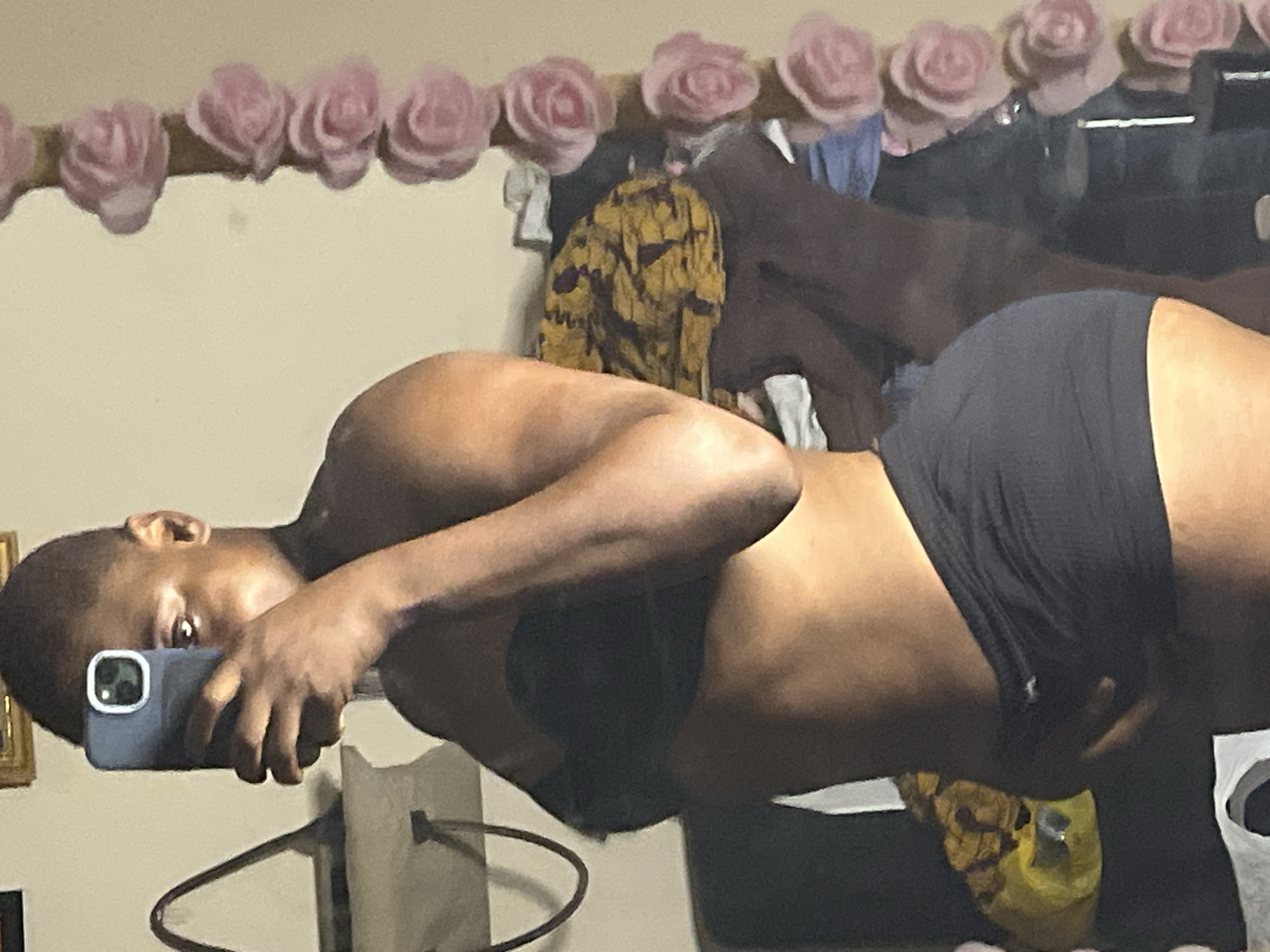 A person is taking a selfie in front of a mirror. The person has dark skin and short hair and is holding a phone with a blue case. They are wearing what appears to be a dark-colored bra and shorts. The background shows a wall with a garland of pink flowers along the top. A coat hanger and some clothing are visible in the reflection of the mirror. It is impossible to determine if this image was taken in Jalingo, Nigeria, with the information available.