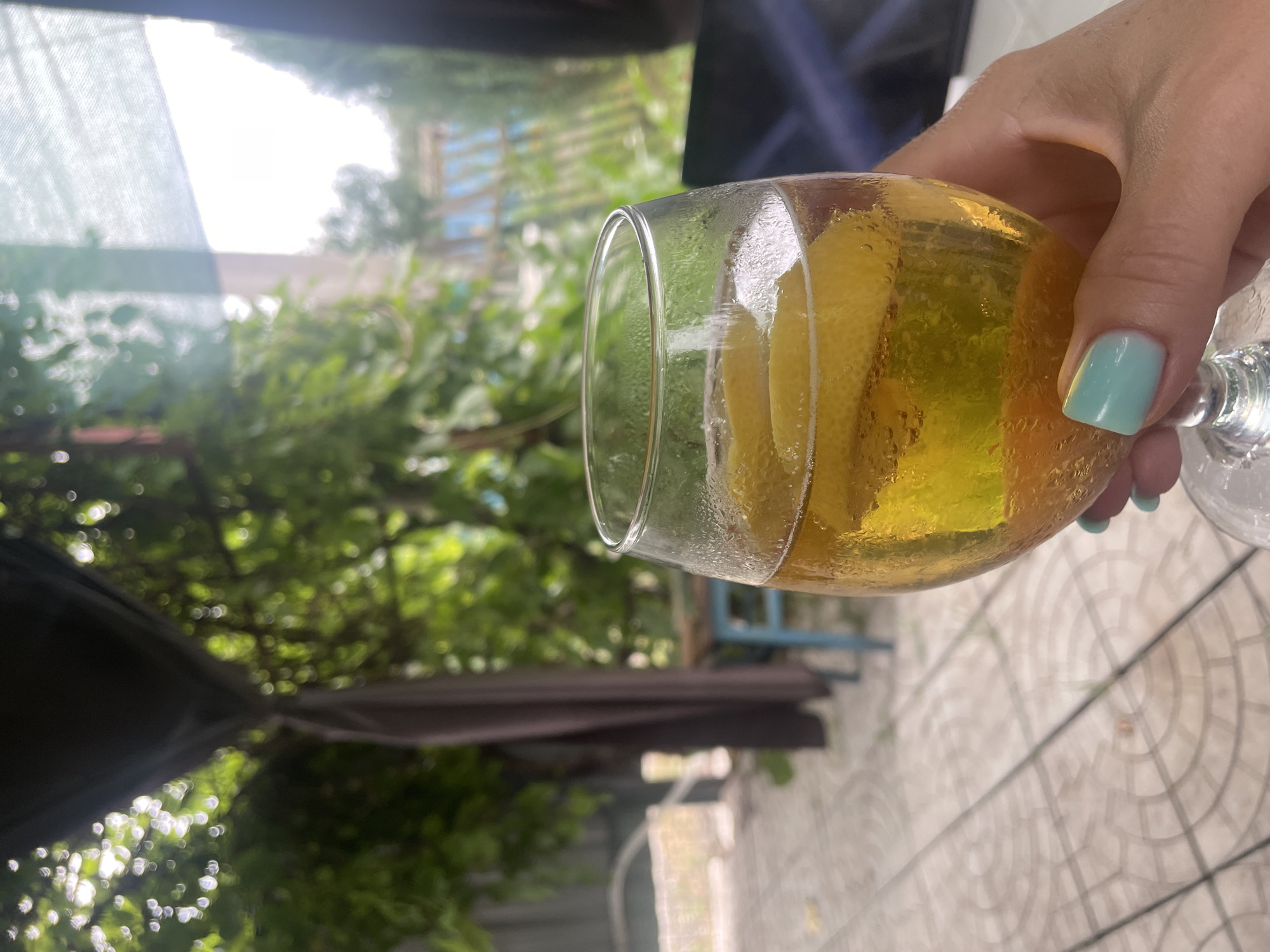 A hand with light blue nail polish holds a glass of amber liquid with lemon slices, suggesting a refreshing beverage. The glass is condensation-covered and filled with bubbles, indicating it is cold. The background is softly blurred, showing lush green foliage, a patio area with circular patterned tiles, and a hint of a fence. The scene appears to be set outdoors, possibly in a yard or garden. The overall impression is one of relaxed outdoor leisure.