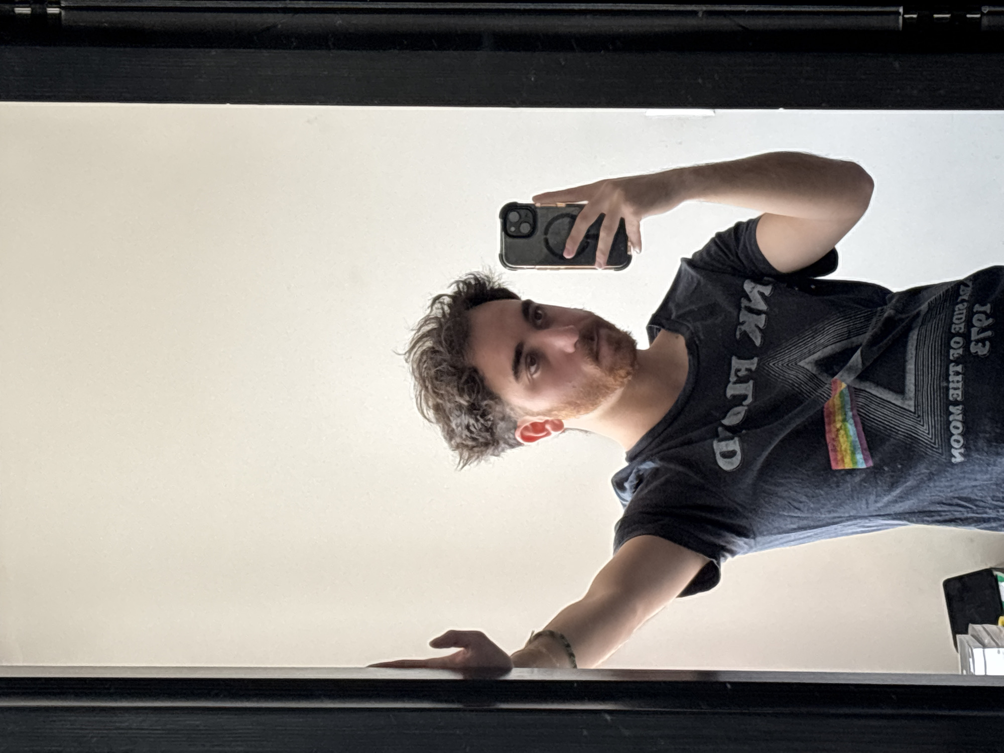 A man is taking a selfie in a mirror. The man has curly brown hair and a reddish-brown beard. He is wearing a dark gray T-shirt with a design on it, possibly of a prism with a rainbow. He's holding a black smartphone, and his arm is extended to take the picture. The background is a plain, light-colored wall. The mirror has a black frame. The location is unspecified, and there's no information to determine if the image was taken in Santa Clara Ocoyucan, Mexico.