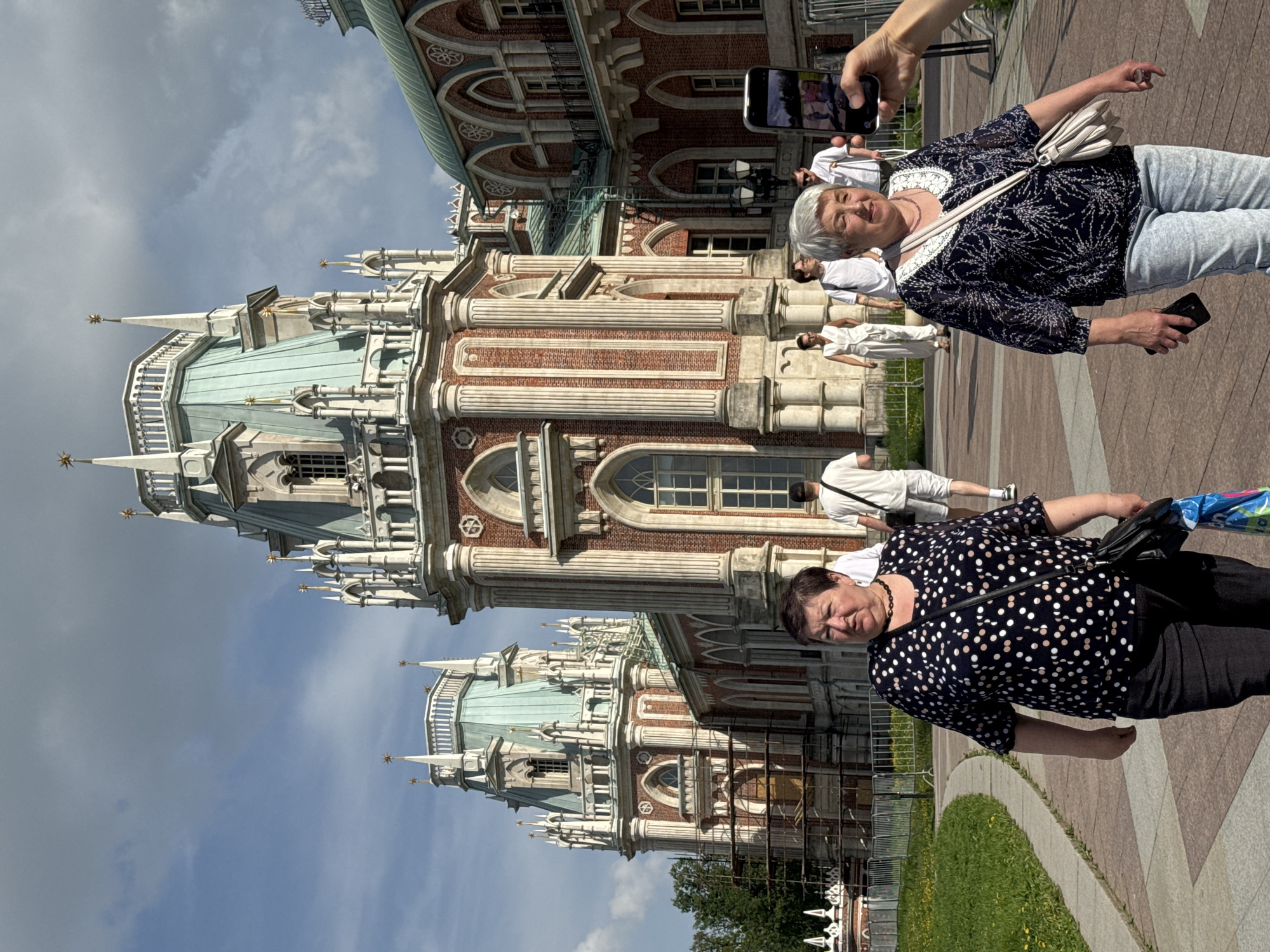 The image shows two women in the foreground, with the Tsaritsyno Palace in Moscow, Russia, in the background. The woman on the right is older, with short grey hair, wearing a dark top with white lace trim, light jeans, and a white shoulder bag. She is holding a black smartphone in her left hand, while a hand holding another phone is taking her photo. The woman on the left has short brown hair and is wearing a black and white polka-dot top and black pants. She is carrying a black shoulder bag and an umbrella. In the background, there are other people, mainly dressed in white, walking along a path in front of the palace. The palace is a large, ornate building with red brick and white stone details, including decorative columns, arches, and spires. The sky is partly cloudy. The ground is paved with a patterned stone.