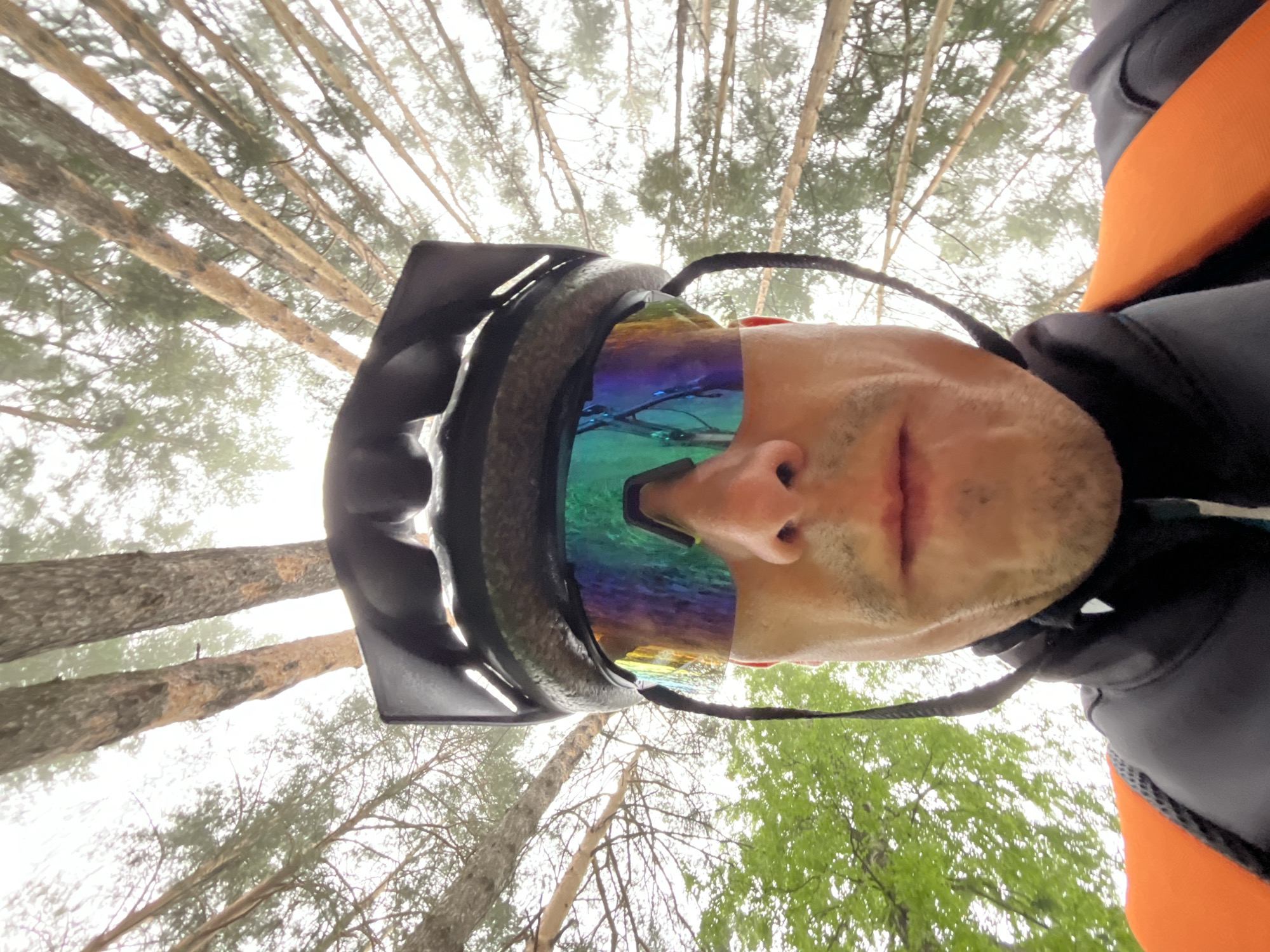 A first-person, inverted perspective captures a person looking upwards into a forest canopy. The main subject is a person, likely male, whose face is partially visible, wearing a dark full-face helmet with large, reflective, iridescent goggles that display hues of green, blue, and purple. A small amount of stubble is visible on their chin and upper lip. They are also wearing a dark top with a bright orange section visible on the right side of the frame, possibly part of a jacket or backpack.
The setting is a dense forest, with numerous tall, slender tree trunks and their branches reaching towards an overcast or foggy sky. Many of the trees appear to be evergreens, possibly pines or firs, with some lighter green foliage from deciduous trees visible towards the bottom right. The diffused light suggests a cloudy day, possibly with mist or fog obscuring the distant sky and tree tops.
The image conveys an immersive outdoor activity, likely from the perspective of someone engaged in mountain biking, cycling, or a similar helmet-wearing sport, given the gear. The upside-down orientation suggests the camera might be mounted on the helmet, capturing the view straight up. The overall atmosphere is one of natural exploration in a temperate forest. No visible text is present in the image. The location is Chaikovsky, Russia.