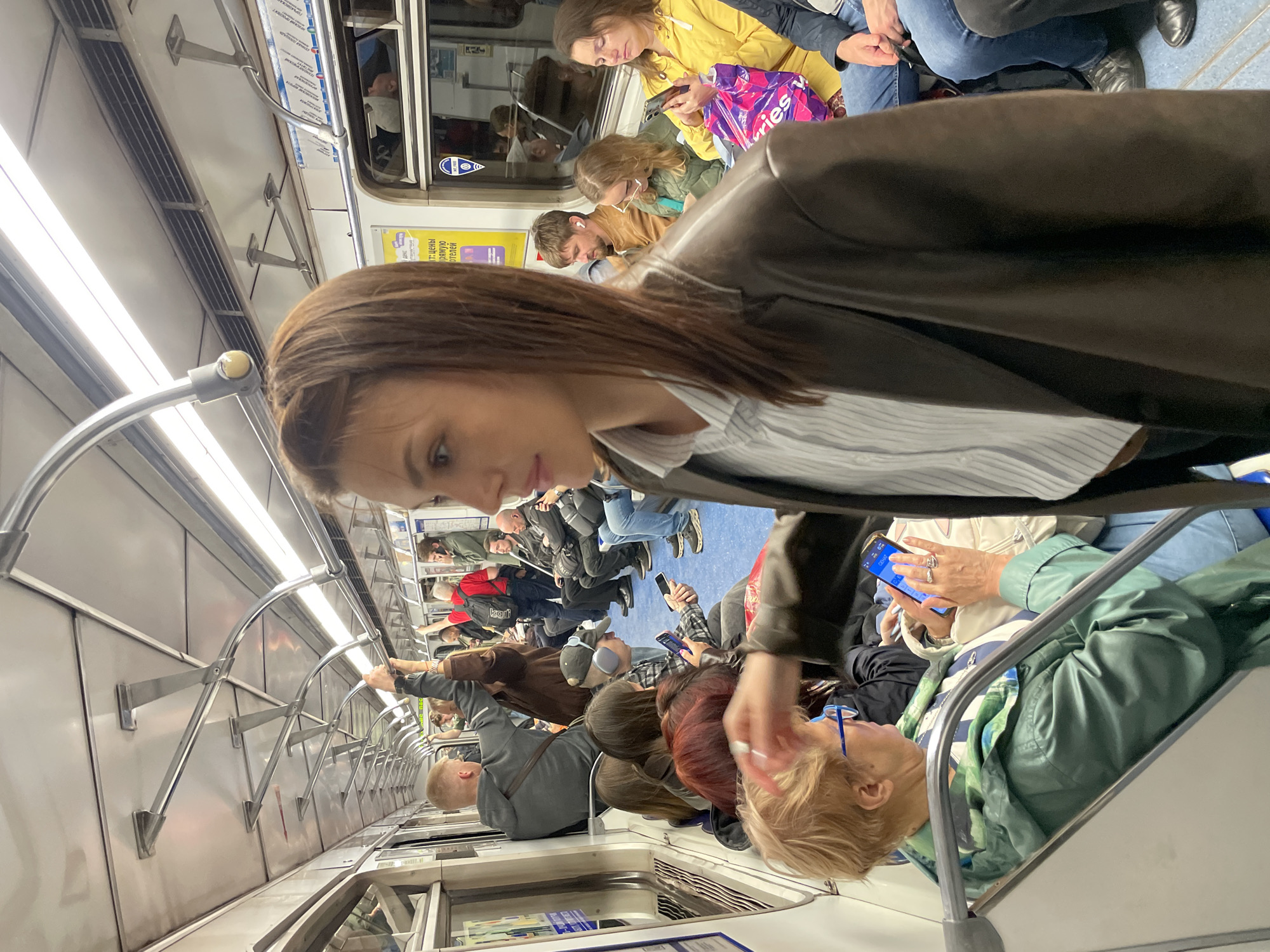 The image shows the interior of a subway car, likely during peak hours due to the number of passengers. Most passengers are standing and holding onto the grab bars overhead. Many people are using their cell phones. The car has gray and white interior panels, and overhead lighting. There is no information that identifies the city in which this image was taken.