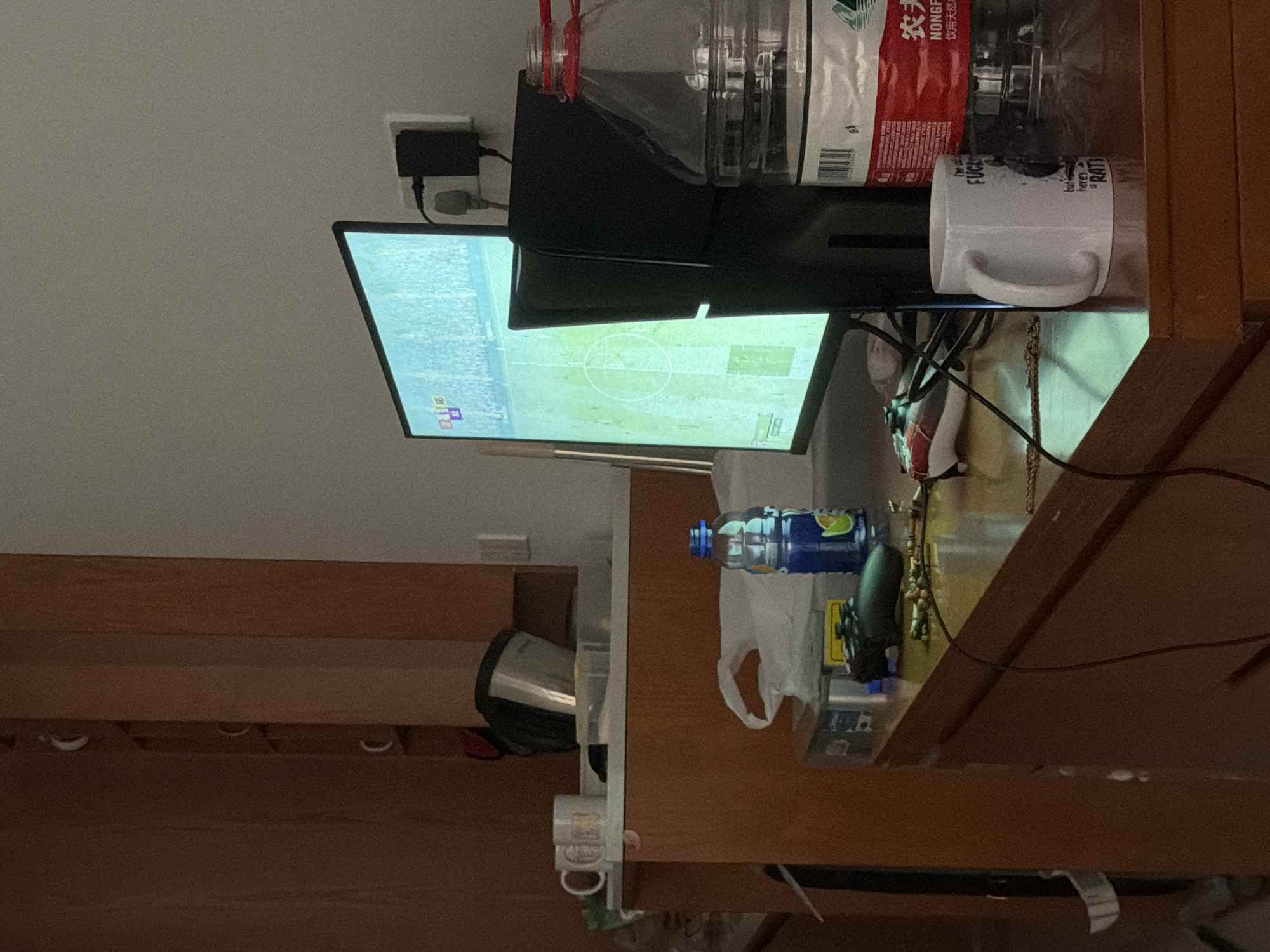 This indoor scene features a computer monitor displaying a soccer game. A black computer tower is positioned behind the monitor. Beside the monitor rests a white mug with bold lettering. A clear plastic bag is perched atop the tower. Beneath the desk, there is an additional mug and a kettle. A gamepad controller and a blue bottle can be seen on the desk. No people are visible. The precise location within Jiangning District, China cannot be determined from this image.