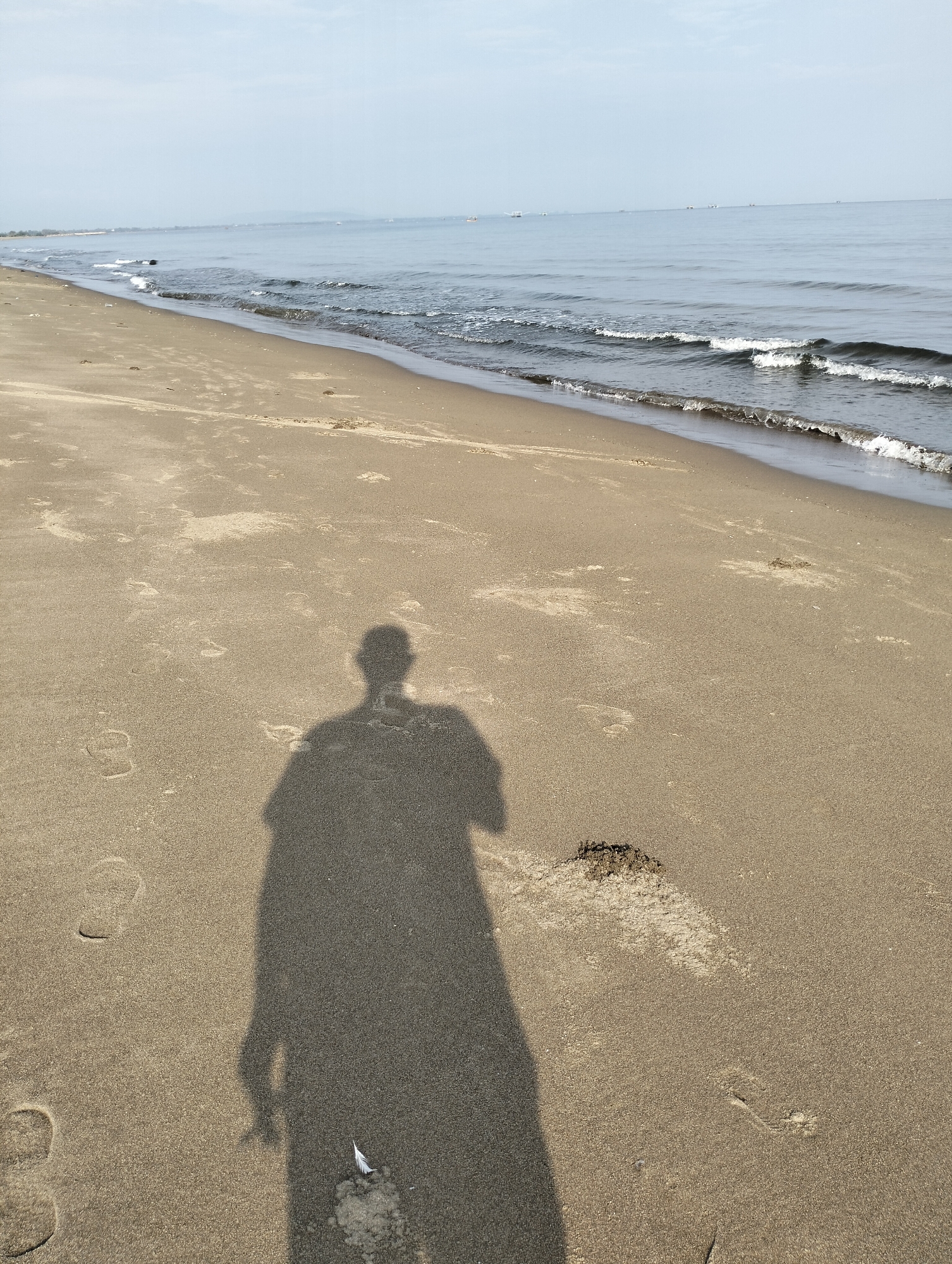 The scene depicts a sandy beach located in Curehtunong, Indonesia. A shadow of a person is cast in the foreground. The beach stretches to the horizon where the ocean meets the sky. Small waves are visible at the shoreline. The sand is marked with footprints and appears moist in some areas. A small, white object (possibly a feather) lies near the base of the shadow. In the distance, there are hints of land and what appear to be boats on the water. The sky is overcast.