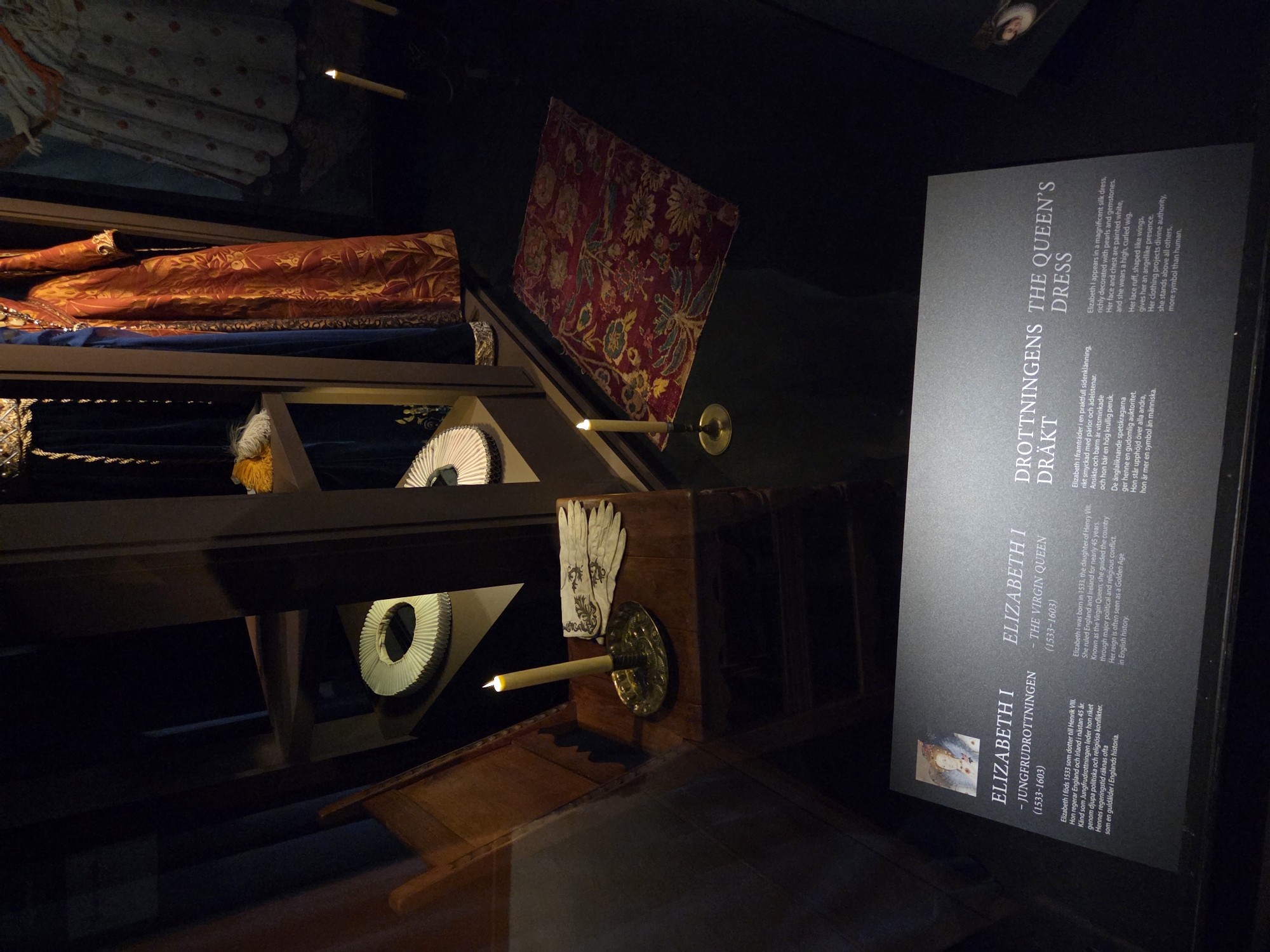 The image depicts an exhibit likely in Malmö, Sweden, showcasing historical artifacts and information related to Queen Elizabeth I. In the foreground, a dark display case contains a wooden chair with a lit candle in a brass holder, placed next to a pair of white gloves. Behind the chair, on shelves, are several white ruffs, commonly worn during that historical period. Further back, on a higher shelf, are two elaborate historical dresses, one a deep blue velvet and the other a richly patterned orange and gold brocade.
Below the exhibit, a dark informational plaque is mounted, featuring portraits and text in both Swedish and English. The Swedish text on the left reads "ELIZABETH I - JUNGFRUDROTTNINGEN (1533-1603)" and describes Elizabeth I as the daughter of Henry VIII, who reigned over England and Ireland for nearly 45 years and is known for her political and religious conflicts, with her reign considered a golden age in English history. The English text on the right reads "ELIZABETH I - THE VIRGIN QUEEN (1533-1603)" and reiterates similar biographical details. To the right of this, another section of the plaque titled "DROTTNINGENS DRÄKT" (Swedish) and "THE QUEEN'S DRESS" (English) describes Elizabeth's attire, noting it is a magnificent silk dress adorned with pearls and gemstones, with a ruffled lace collar, a curved wig, and an angelic presence.
The overall environment is dimly lit, with spotlights highlighting the artifacts and the informational display, suggesting a museum or historical exhibition setting. The dark background and strategic lighting create a dramatic and immersive experience for the viewer.