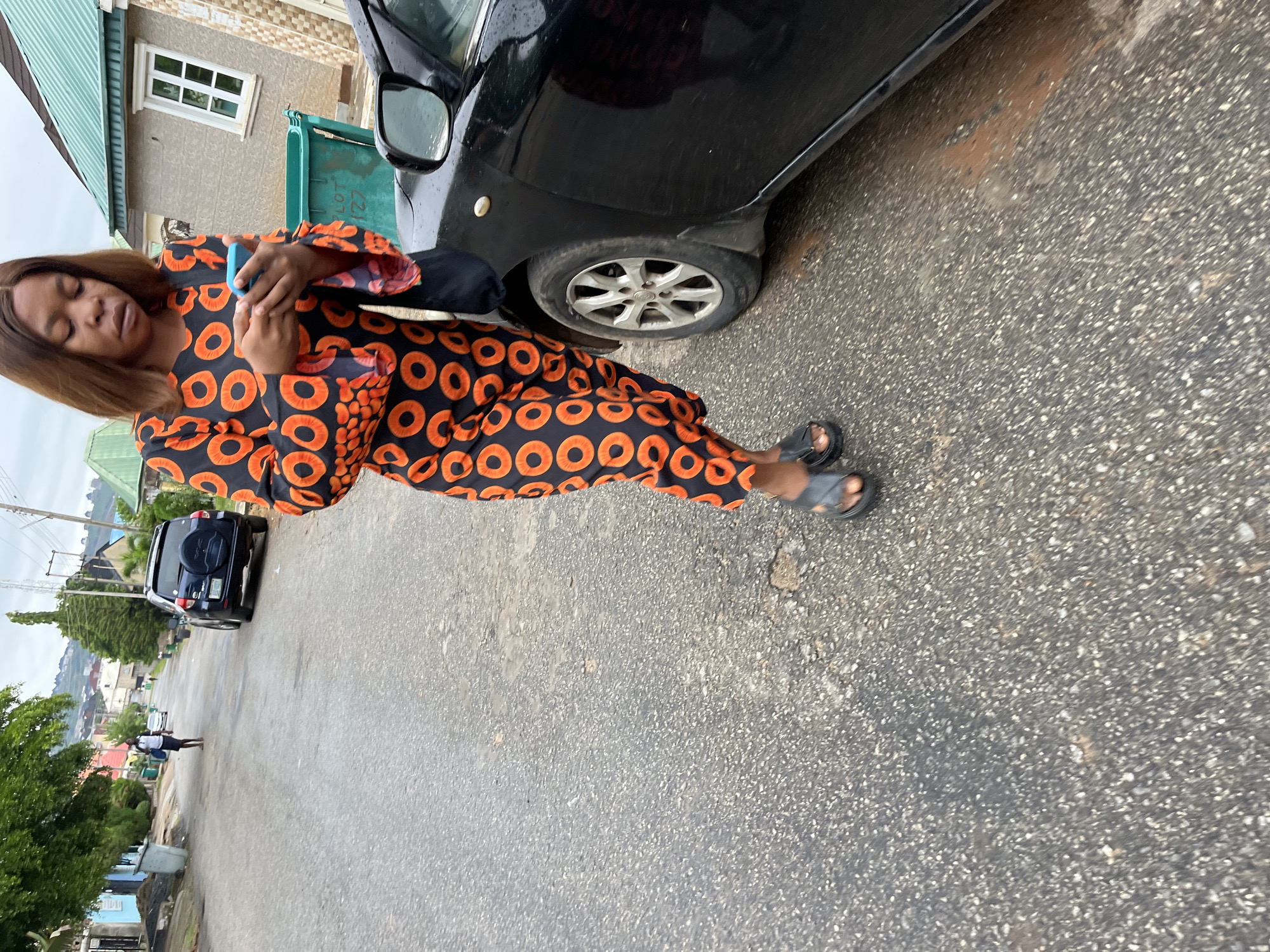 An image captures a woman standing on an asphalt road in what appears to be a residential area in Abuja, Nigeria. The image is taken from a slightly low angle, looking up towards the woman and the street.
The main subject is a woman with brown hair, wearing a long-sleeved dress with a distinctive orange circular pattern on a black background. She is holding a light blue mobile phone with both hands, her gaze directed downwards, suggesting she is focused on the device. Her eyes appear to be closed or cast down. She is wearing dark sandals on her feet.
To her right, a black car is parked partially visible, with its front wheel and side mirror in view. Behind the woman and the car, there's a building with light brown walls and a white-framed window. A green object, possibly a waste bin or utility box, is situated near the car, bearing the text "LOT 127".
Further down the street, another black SUV or car is parked. In the distance, a smaller figure, possibly a child or another person, is visible walking on the road. The road itself is paved with asphalt, showing some wear. The background features more buildings, green trees, and distant hills under an overcast sky, indicating daytime.