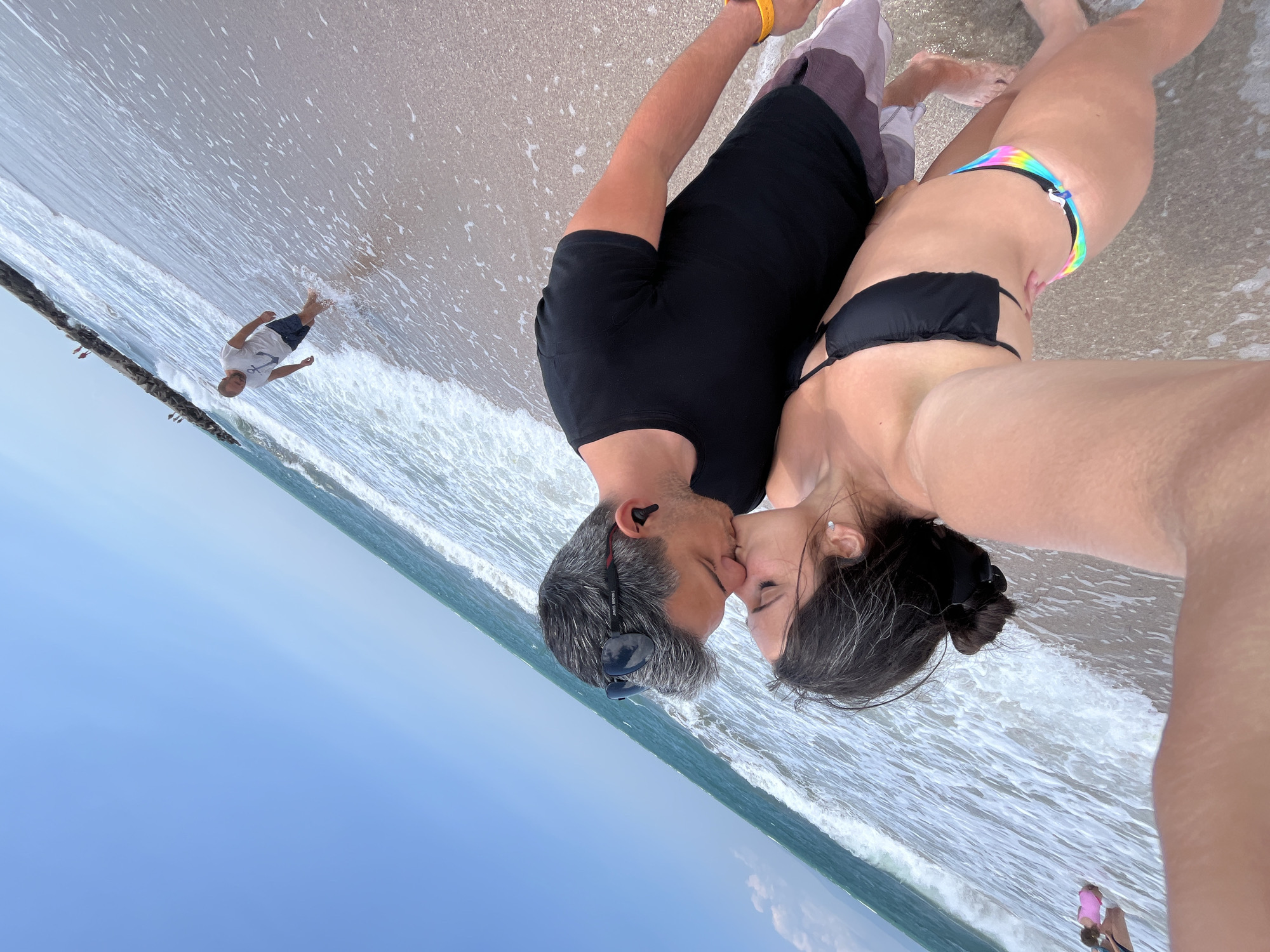In Pomorie, Bulgaria, a couple is captured in a close-up selfie on the beach, sharing a kiss as waves gently lap at their feet. The man, wearing a black t-shirt and patterned shorts, has sunglasses perched on his head and is wearing black earbuds. The woman, in a black bikini, has her hair pulled back and is wearing a small earring. In the background, a man in a white t-shirt with an anchor design and dark shorts stands in the shallow water, further down the beach. A stone jetty extends into the sea, with several figures visible at its end, and the vast expanse of the sky and ocean completes the scene.