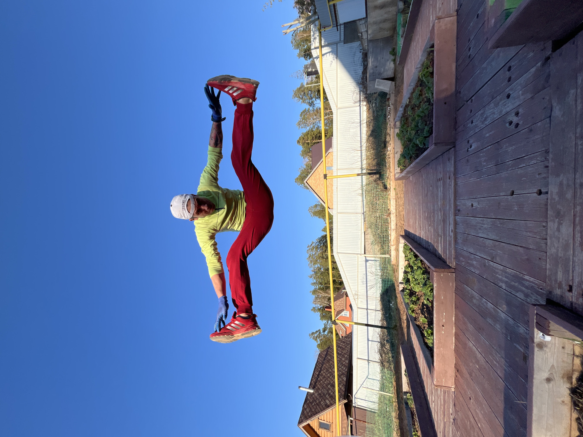 The image, oriented with the sky at the bottom and the ground at the top, shows a person stretching. The person is wearing red pants, a light green long-sleeved shirt, red sneakers, a white cap, and blue gloves. They are bending over backward, holding their feet. The background includes a wooden deck with built-in planters, a white fence, several buildings with metal or shingled roofs, trees, and a clear blue sky. This scene is located in Chaikovsky, Russia.