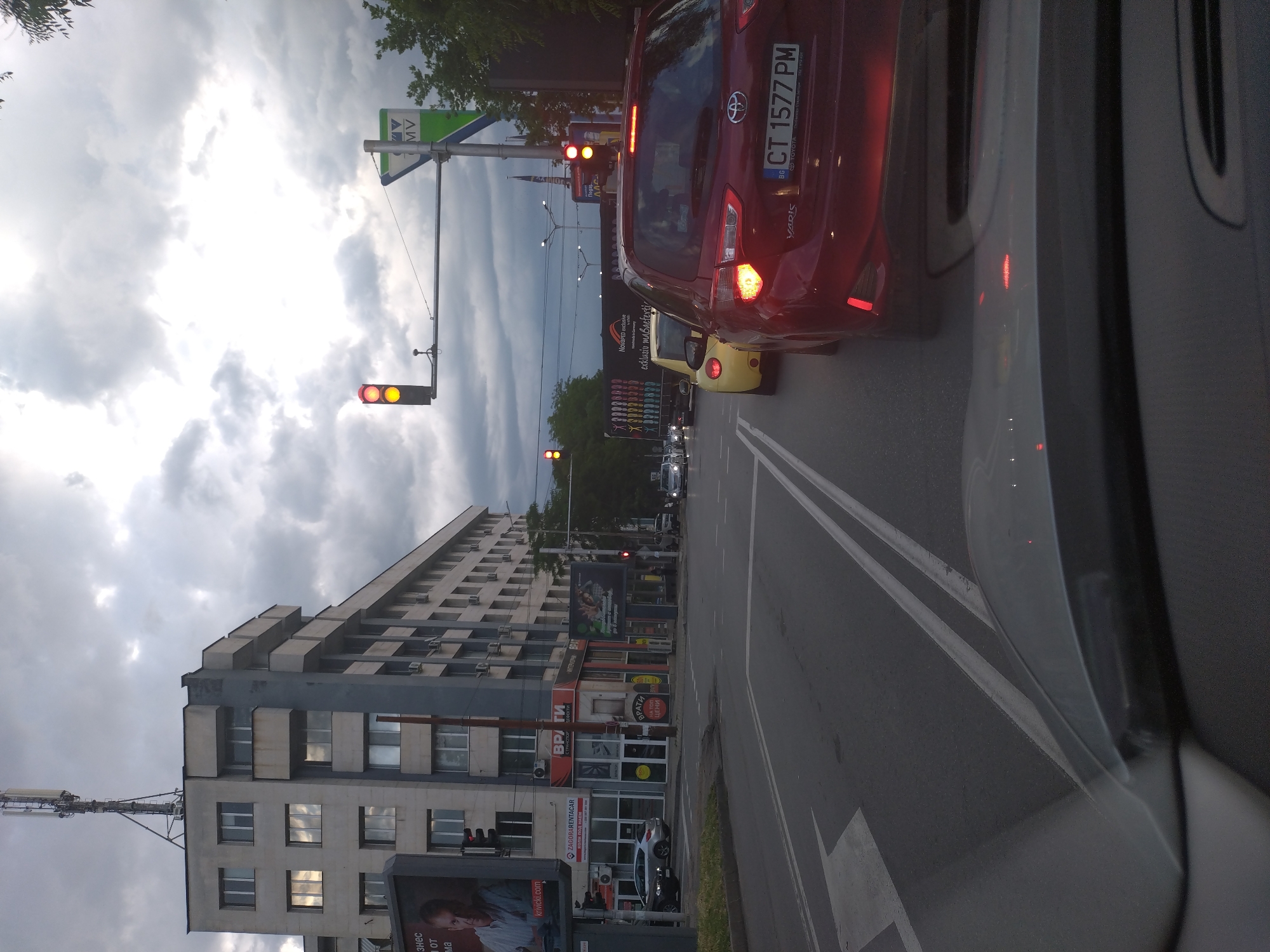 A daytime image, likely taken from inside a vehicle, captures a street scene in an urban environment, specifically Stara Zagora, Bulgaria. The sky is overcast with heavy, dark clouds.
In the foreground, the edge of a car's dashboard or window frame is visible on the right, suggesting the perspective of a driver or passenger. The road ahead is paved, with white lane markings indicating an arrow pointing straight ahead and a double solid line separating lanes.
Traffic is stopped, with a red Toyota Yaris, license plate "CT 1577 PM" (CT indicating Stara Zagora, BG for Bulgaria), positioned in front of the camera car. The car's brake lights are illuminated. Further ahead, a yellow car and other vehicles are visible, also stopped. Traffic lights are prominent; one directly above the red Toyota shows a red signal, and another further down the street also displays red. A green street sign with "MV" and "Под VV" (likely part of a larger sign) is visible near the first traffic light.
To the left, a tall, multi-story building with a somewhat Brutalist architectural style dominates the frame. It has many windows and external air conditioning units. A communications mast with antennas is affixed to its roof. Along the base of this building are several storefronts.
Visible text on various signs and billboards includes:
* Car license plate: "CT 1577 PM", "BG", "TOYOTA", "YARIS".
* On billboards: "знес", "OT", "ма", "krivicki.com", "NovaPED exclusiv", "exklusiv mabaeferti", "ВРАТИ НА ТОП ЦЕНИ" (Doors at Top Prices).
* On storefronts: "ZAGORA RENTACAR", "КОЛИ ПОД НАЕМ" (Cars for Rent), "ВРАТИ", "ТРАНСПОРТ", "ДЕФЕКТИ", "ИНТЕРИОРНИ ВРАТИ" (Doors, Transport, Defects, Interior Doors).
The scene depicts a typical moment of urban traffic, with cars stopped at an intersection under a cloudy sky in Stara Zagora.