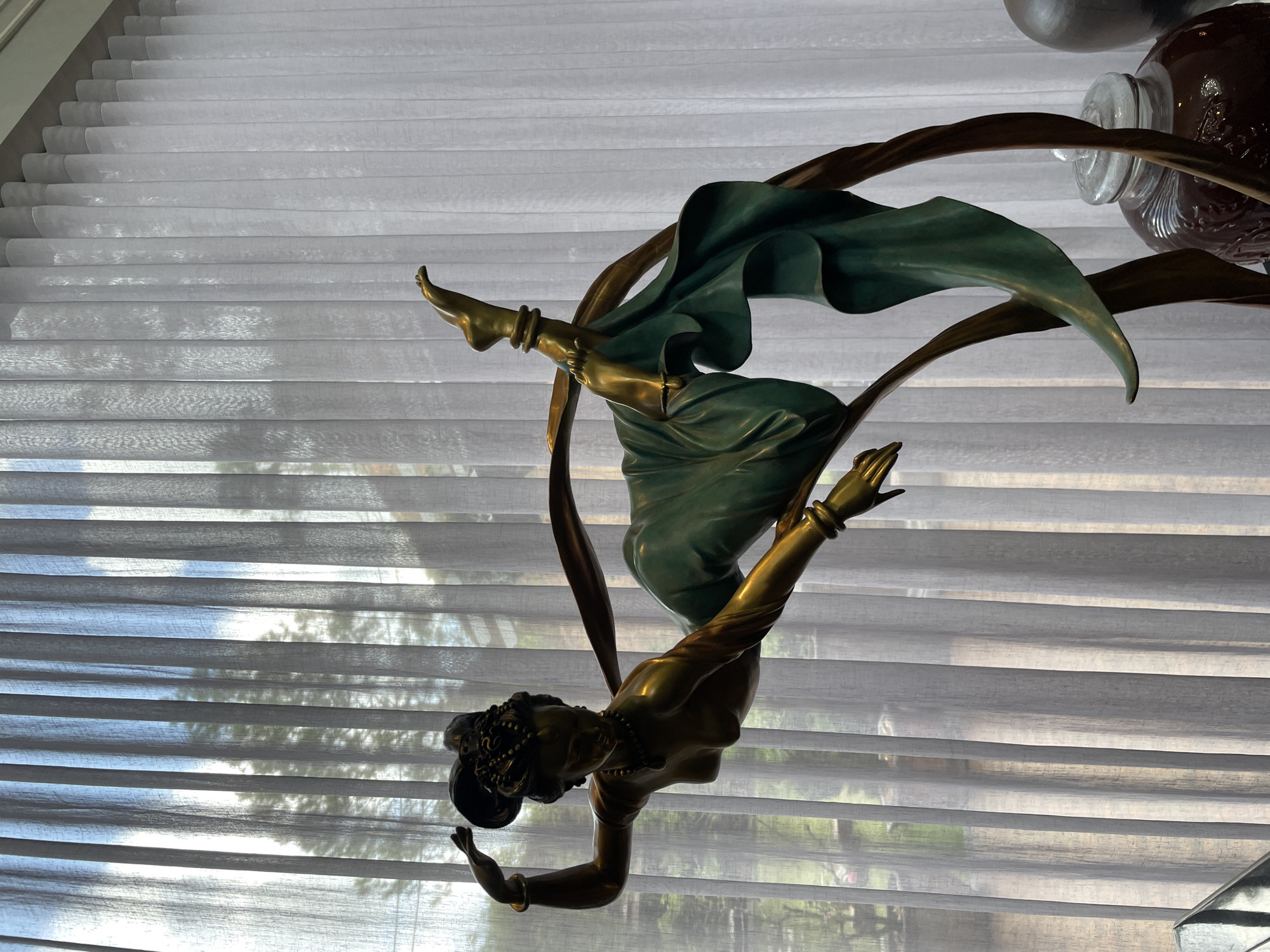 A bronze sculpture of a dancer is positioned in front of sheer white vertical blinds. The dancer is depicted in a dynamic pose, with one leg extended upwards and the other bent. She is adorned with jewelry, including bracelets, necklaces, and an elaborate headdress. A flowing green fabric drapes around her body and trails behind her. The background, visible through the blinds, suggests an outdoor setting with trees and possibly buildings. Below the sculpture, a large glass jar with a dark liquid and a glass lid is partially visible, along with another dark, rounded object. The lighting suggests it is daytime, with sunlight filtering through the blinds. The overall impression is of an artistic display within an interior space overlooking a blurred outdoor environment. The location is identified as Xuân Trường - Đà Lạt, Vietnam.